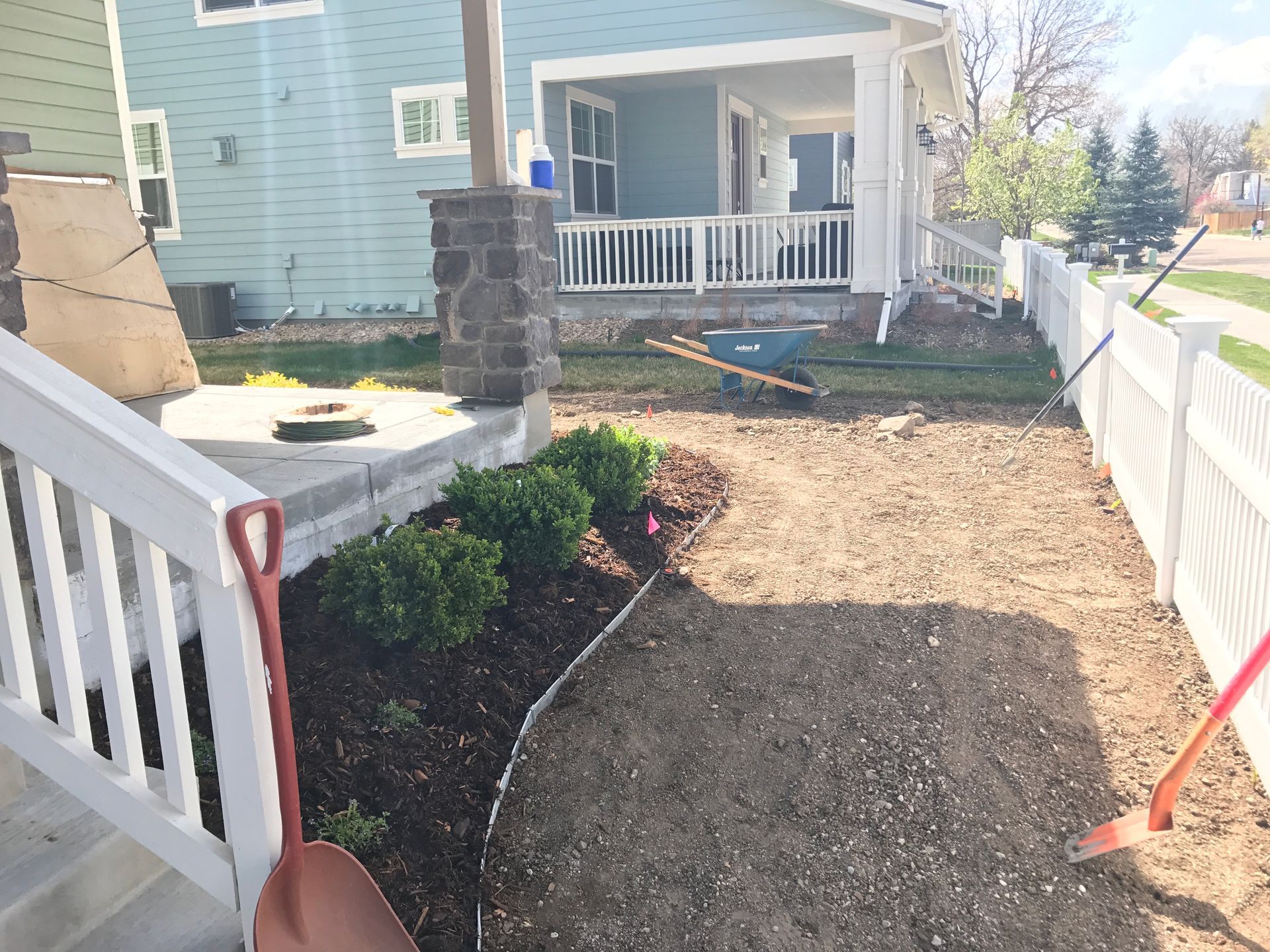Side yard landscaping: gravel path, shrubs, white fence, house with porch. Shovel resting on the path.