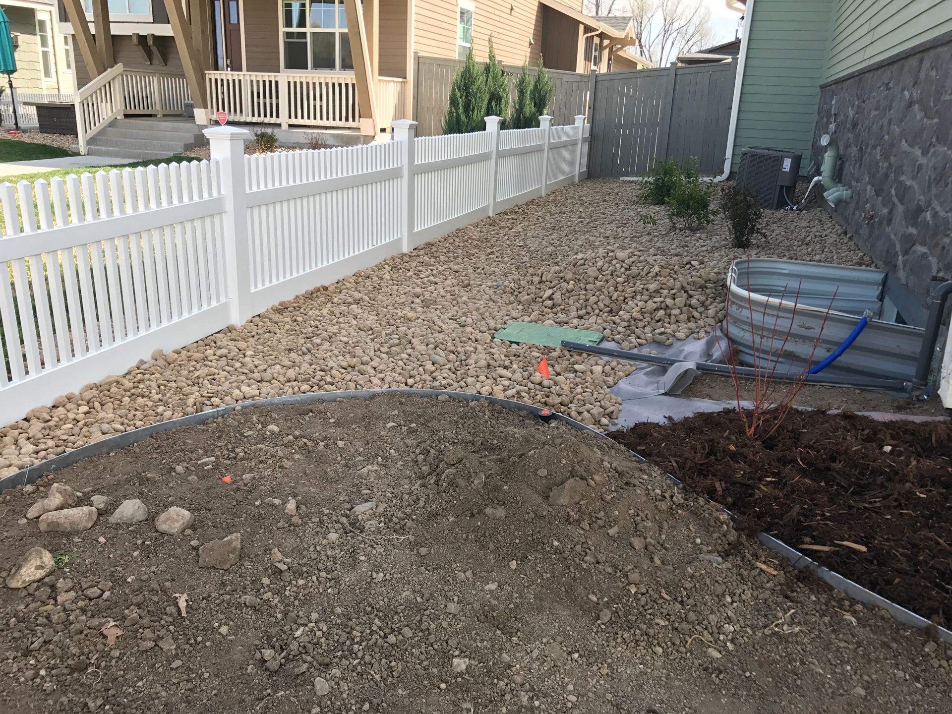 Backyard landscaping with white picket fence, gravel, soil pile, and building.