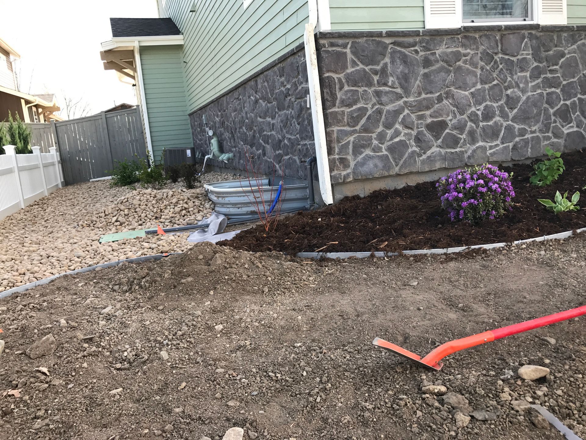 A garden bed with a stone wall and plants, a rake on the dirt ground, a house in the background.