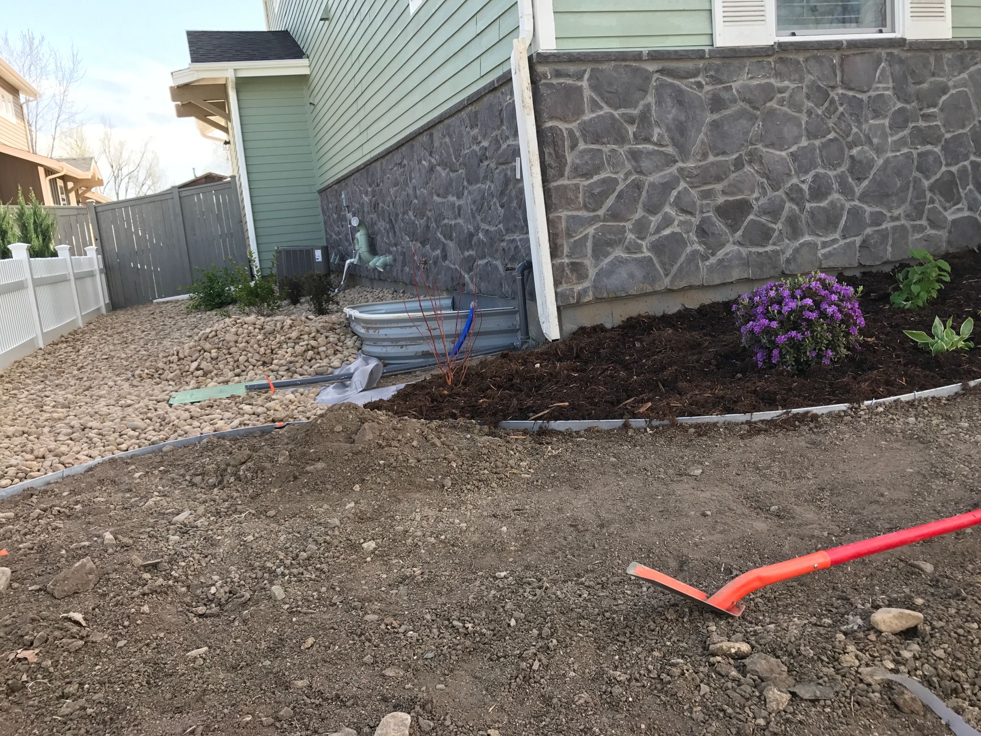 Yard under construction with a metal basin, gravel bed, and a house with green siding and stone foundation.