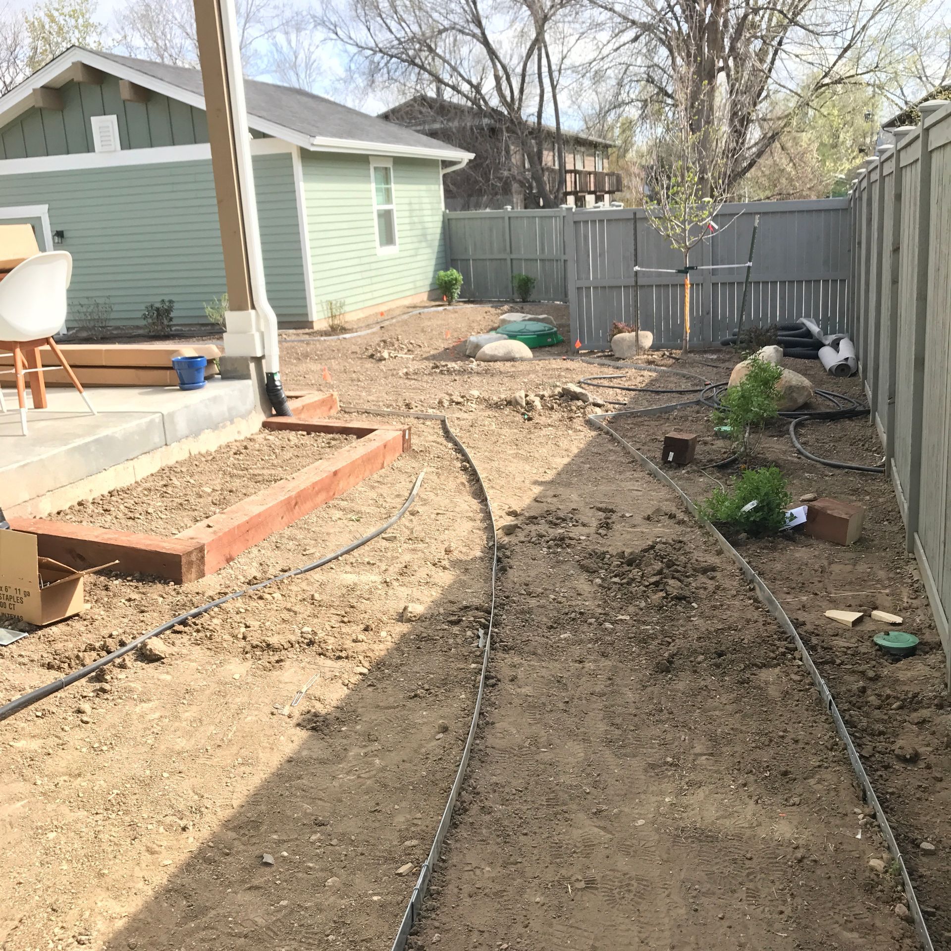 Backyard with garden beds and dirt paths, irrigation system, gray fence and green house.
