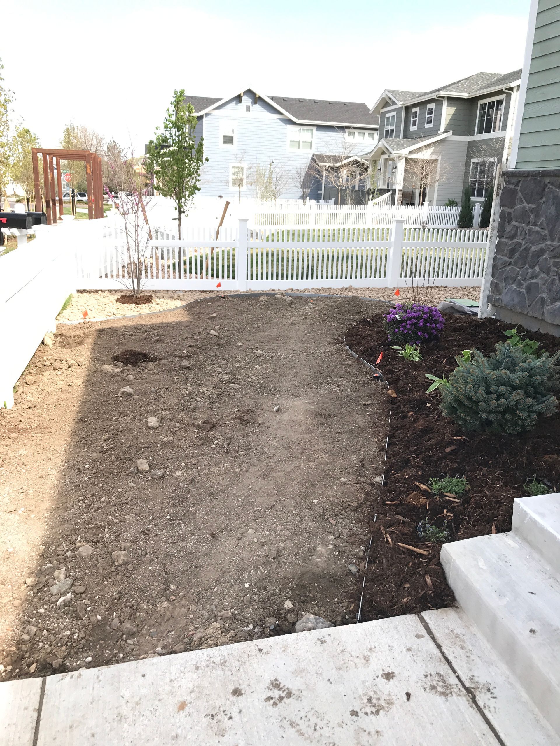 Empty front yard bed with a white picket fence, houses in the background, concrete sidewalk on the left.
