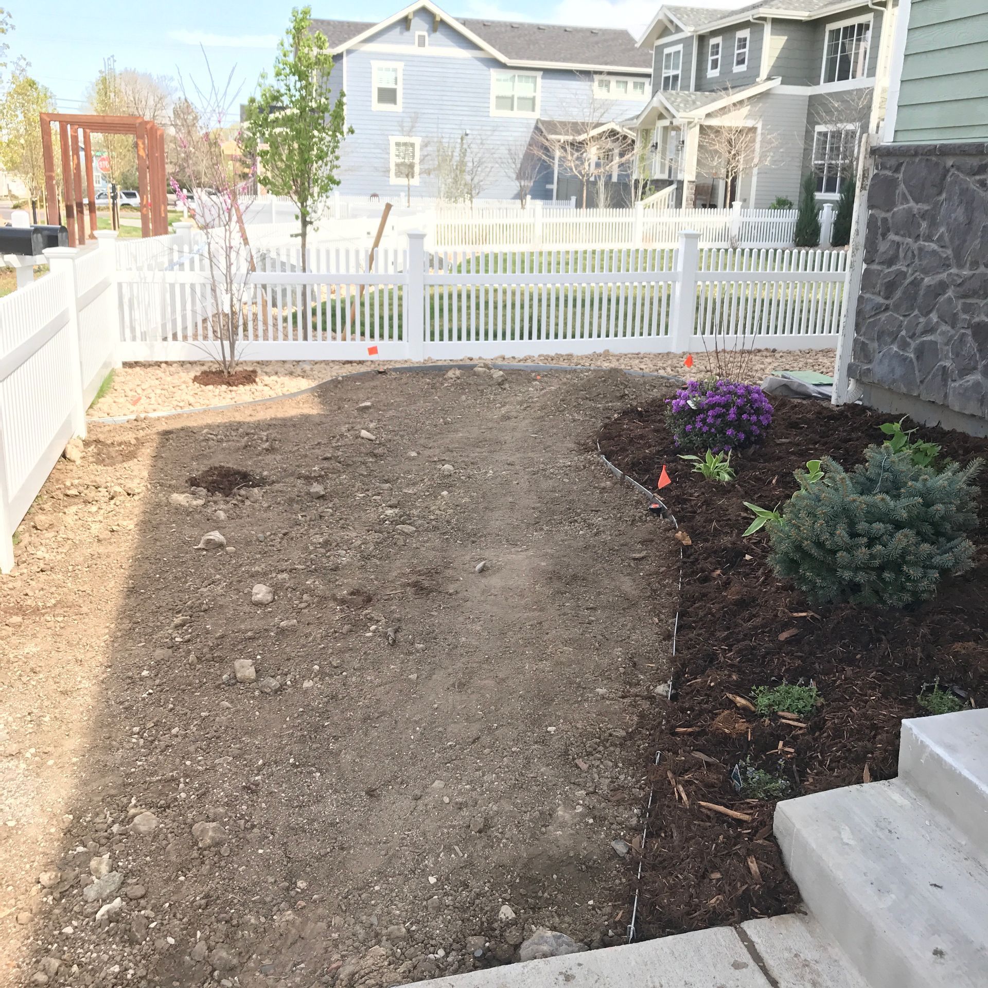 A front yard with white picket fence, dirt, mulch, and greenery; houses in the background.