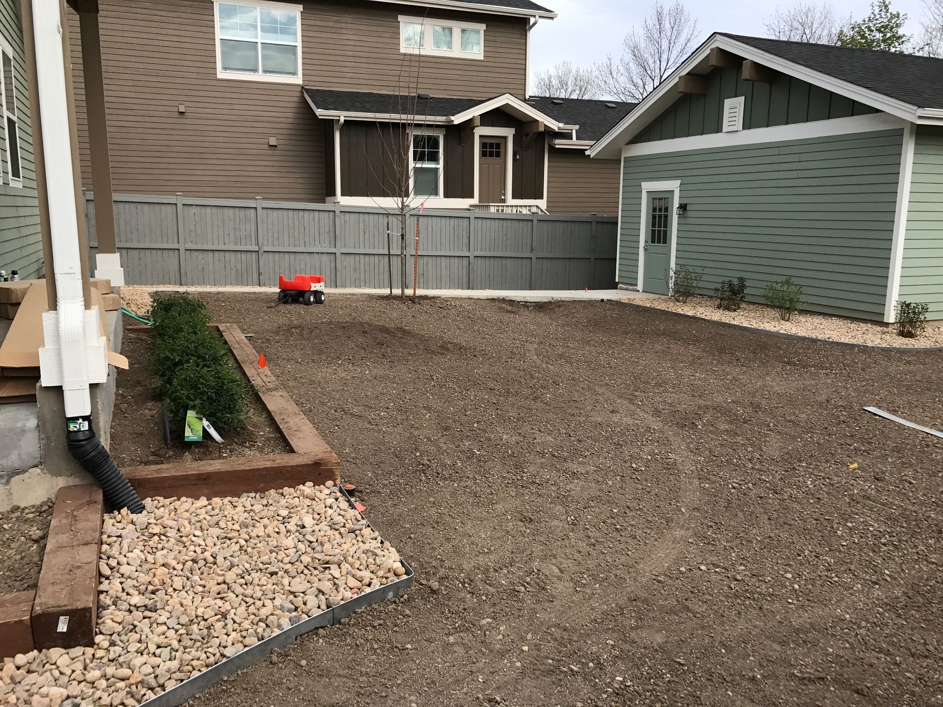 Gravel-covered backyard with low retaining walls, a small red toy, and a few shrubs. Houses and a fence are in the background.