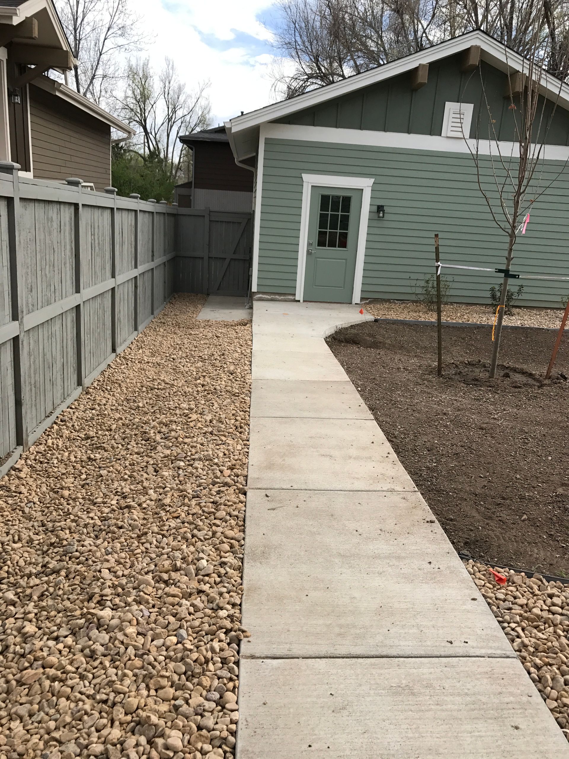 Concrete path leading to a green building with a door, next to a rock bed and a gray fence.