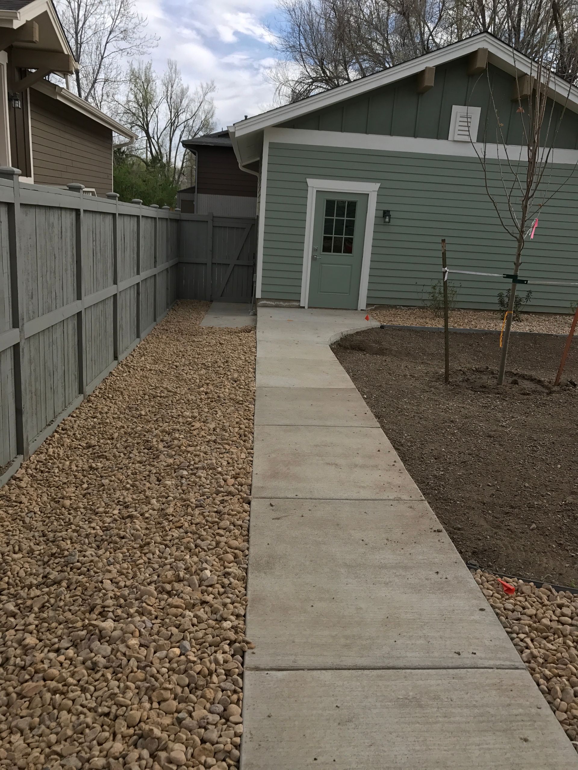 Concrete path leading to a green building with a side yard of gravel and a gray fence.