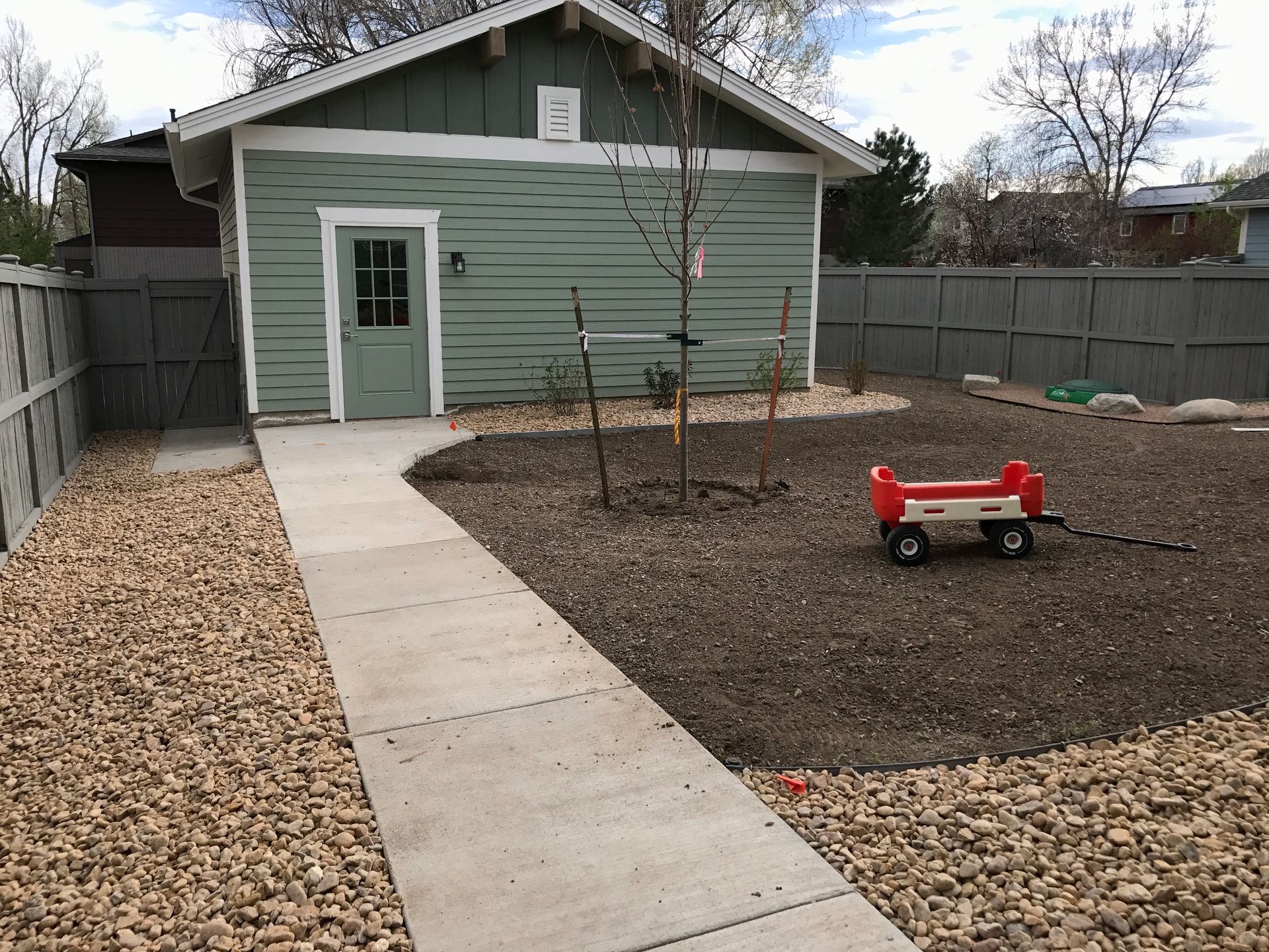 Backyard with a cement path leading to a green building. A red wagon sits in the gravel yard.