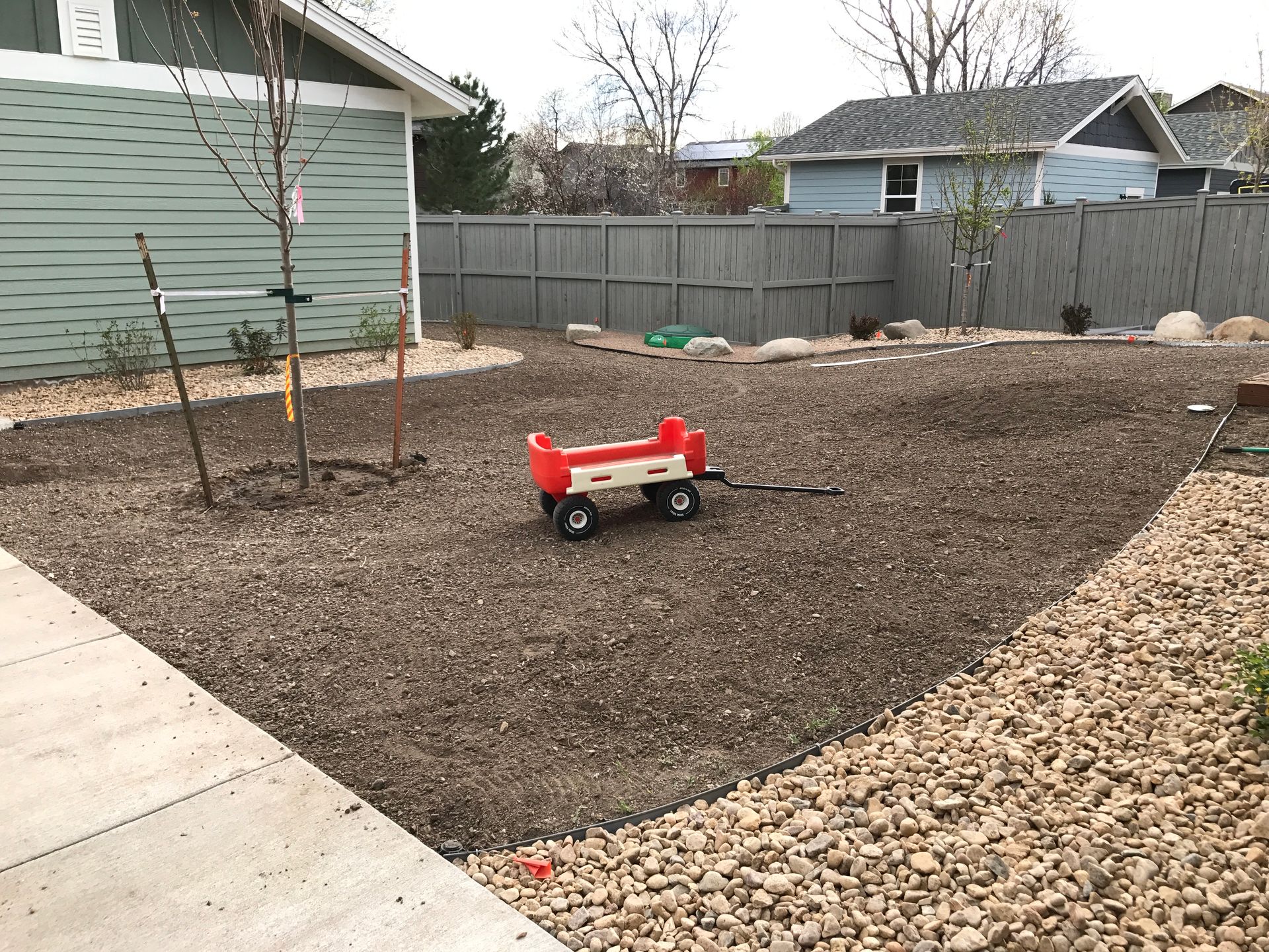 Red wagon in a backyard covered with brown mulch, edged with rocks, next to a concrete path and fence.