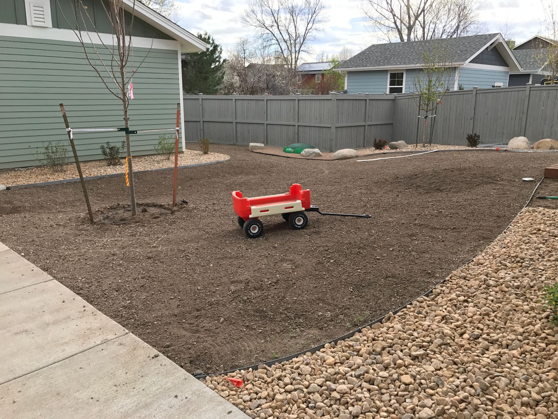 Red wagon in a gravel-covered yard; light blue houses and a gray fence in the background.