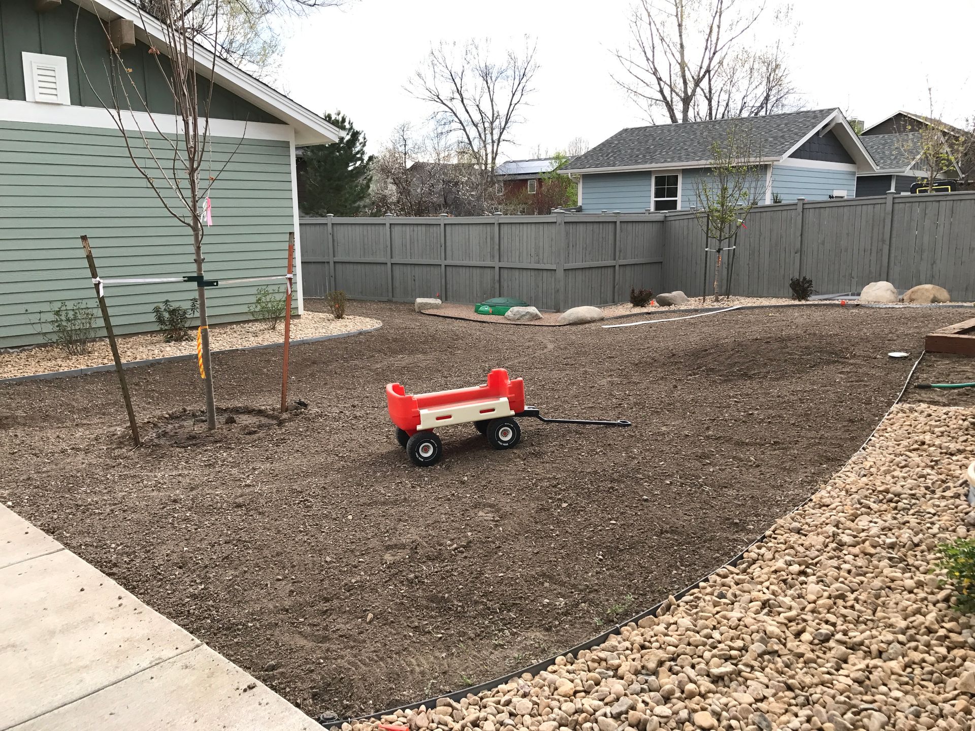 A red and white wagon sits in a gravel backyard bordered by a gray fence and a walkway.