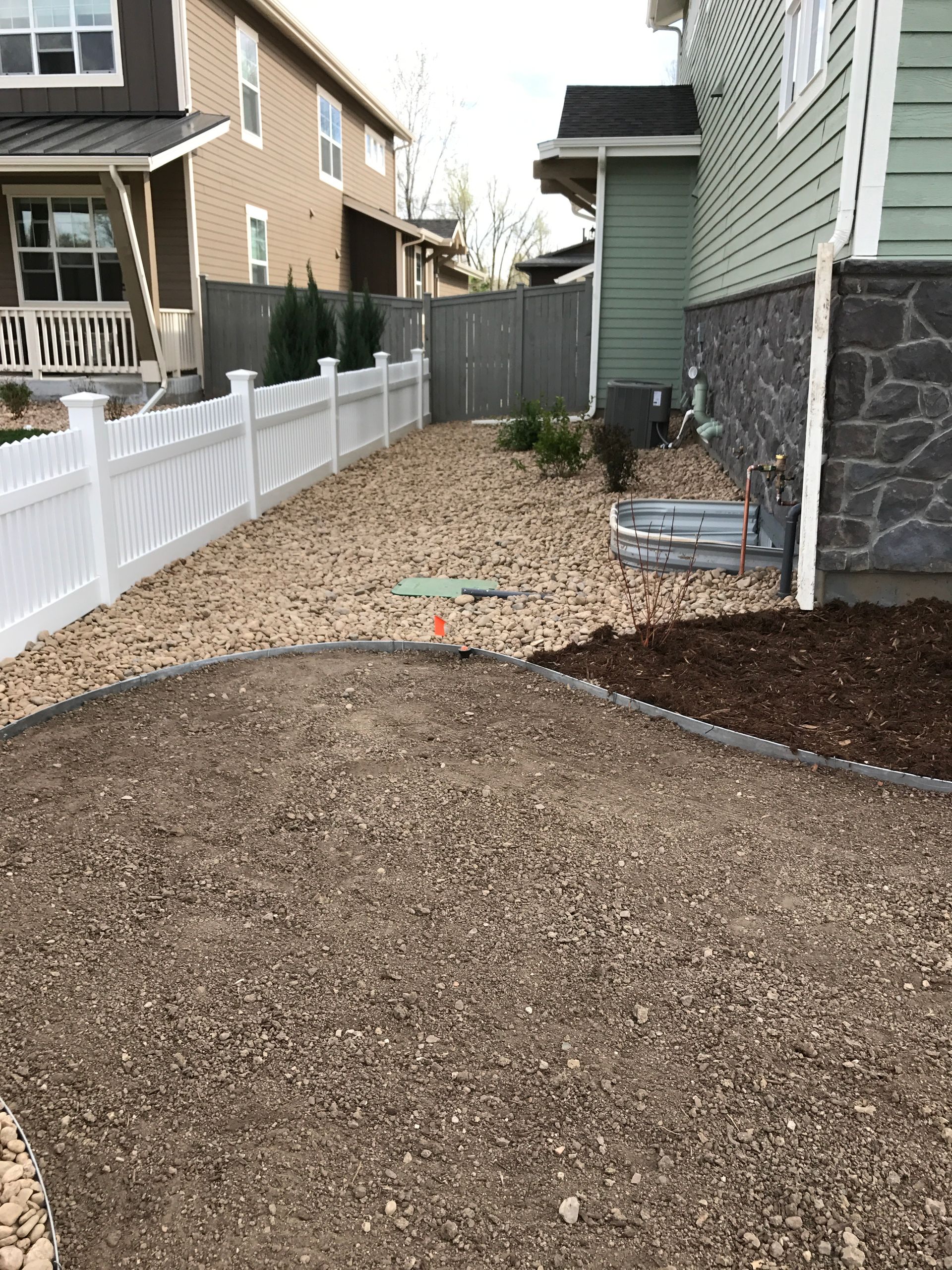 A residential side yard with gravel, white fence, and green house; brown, tan, and gray colors.