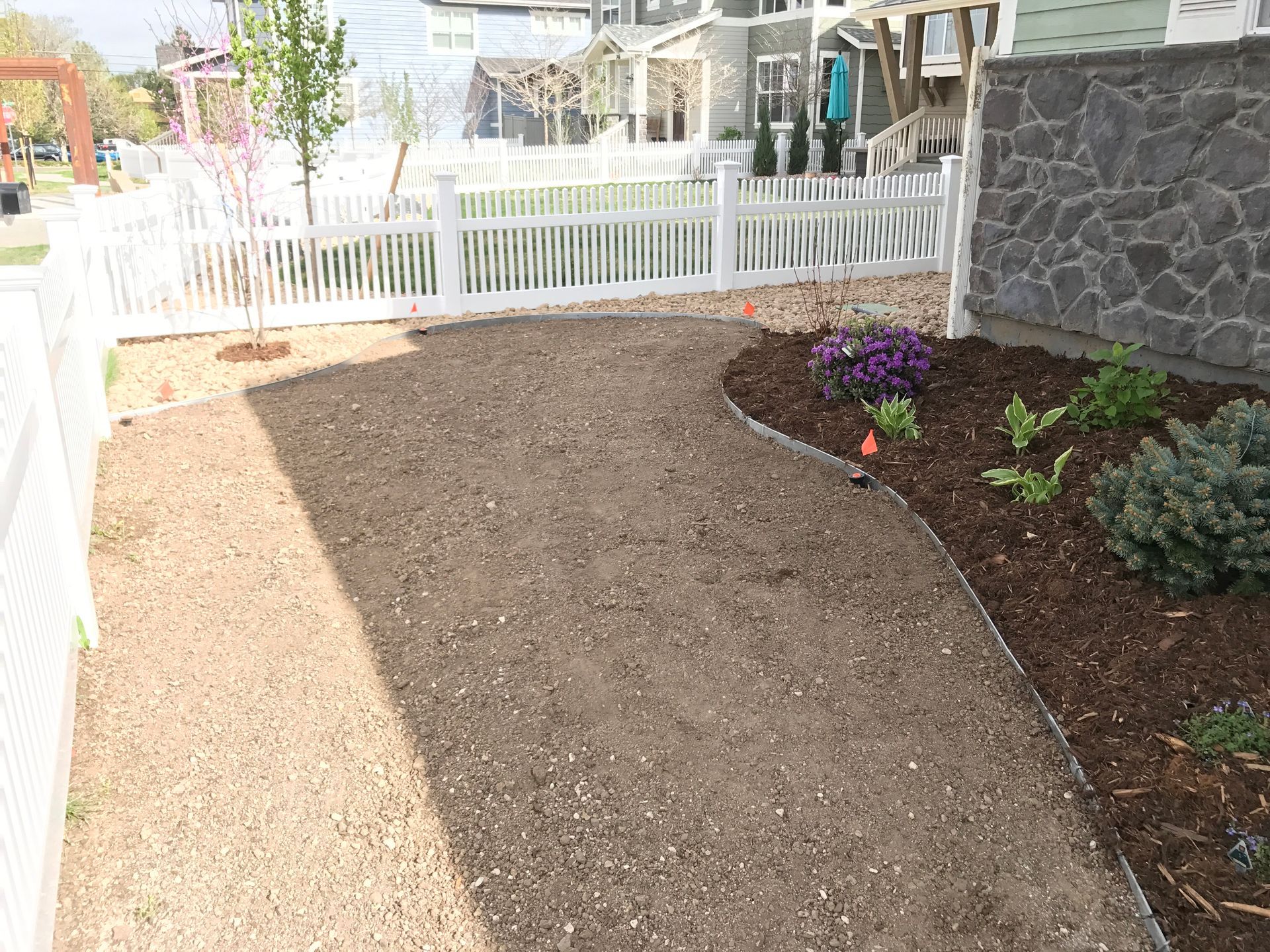 Gravel path curves through a yard bordered by white picket fence and a flower bed with mulch.