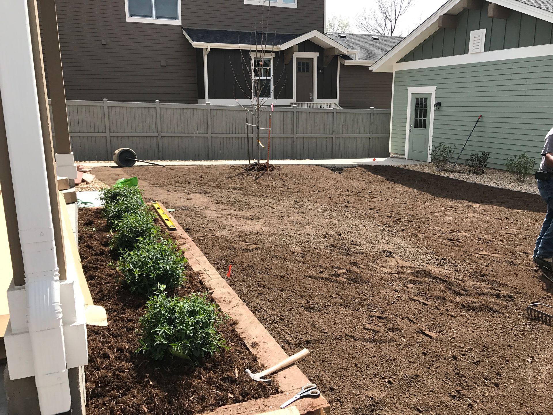 A newly landscaped backyard with mulch, shrubs, and a small tree, next to a house with a green garage.