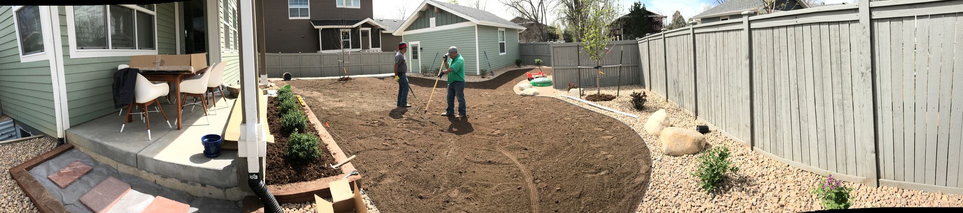 Two people in a backyard. A green house and a gray fence frame the space. The yard is mostly dirt.