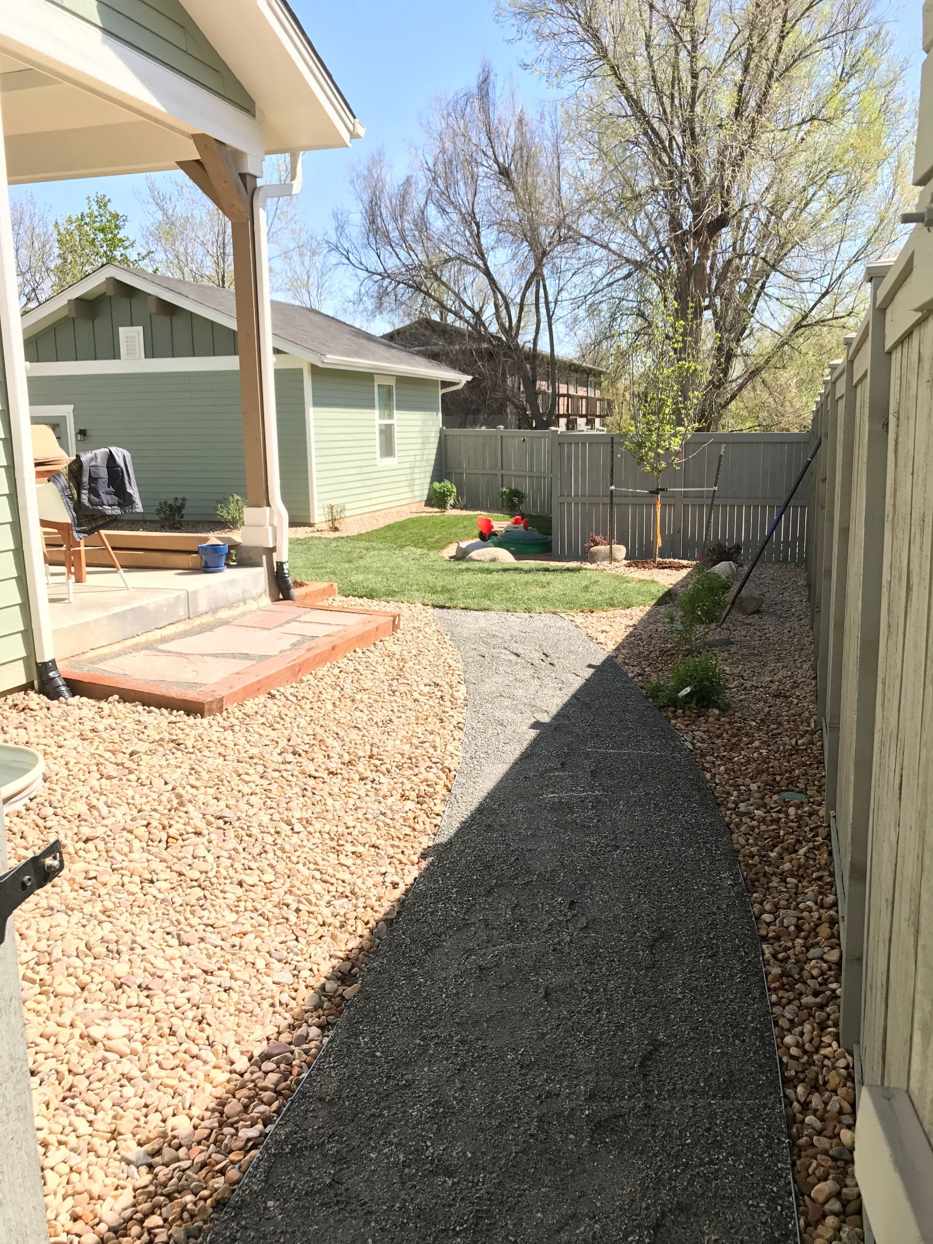 Backyard with gravel path, tan stones, green grass, and fence. A small house is in the background.