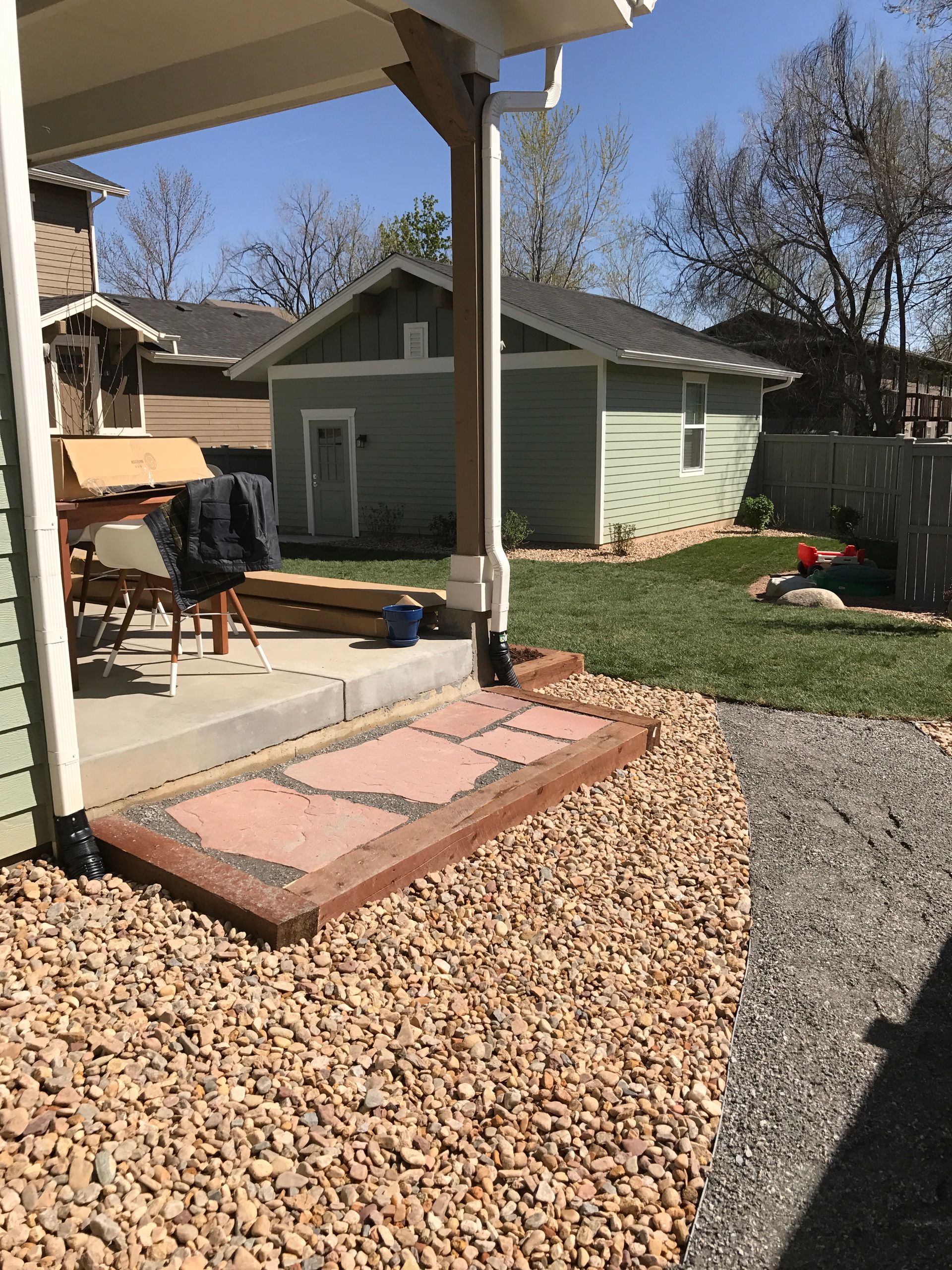 Patio with reddish pavers, tan gravel, green grass, and a green outbuilding under a blue sky.