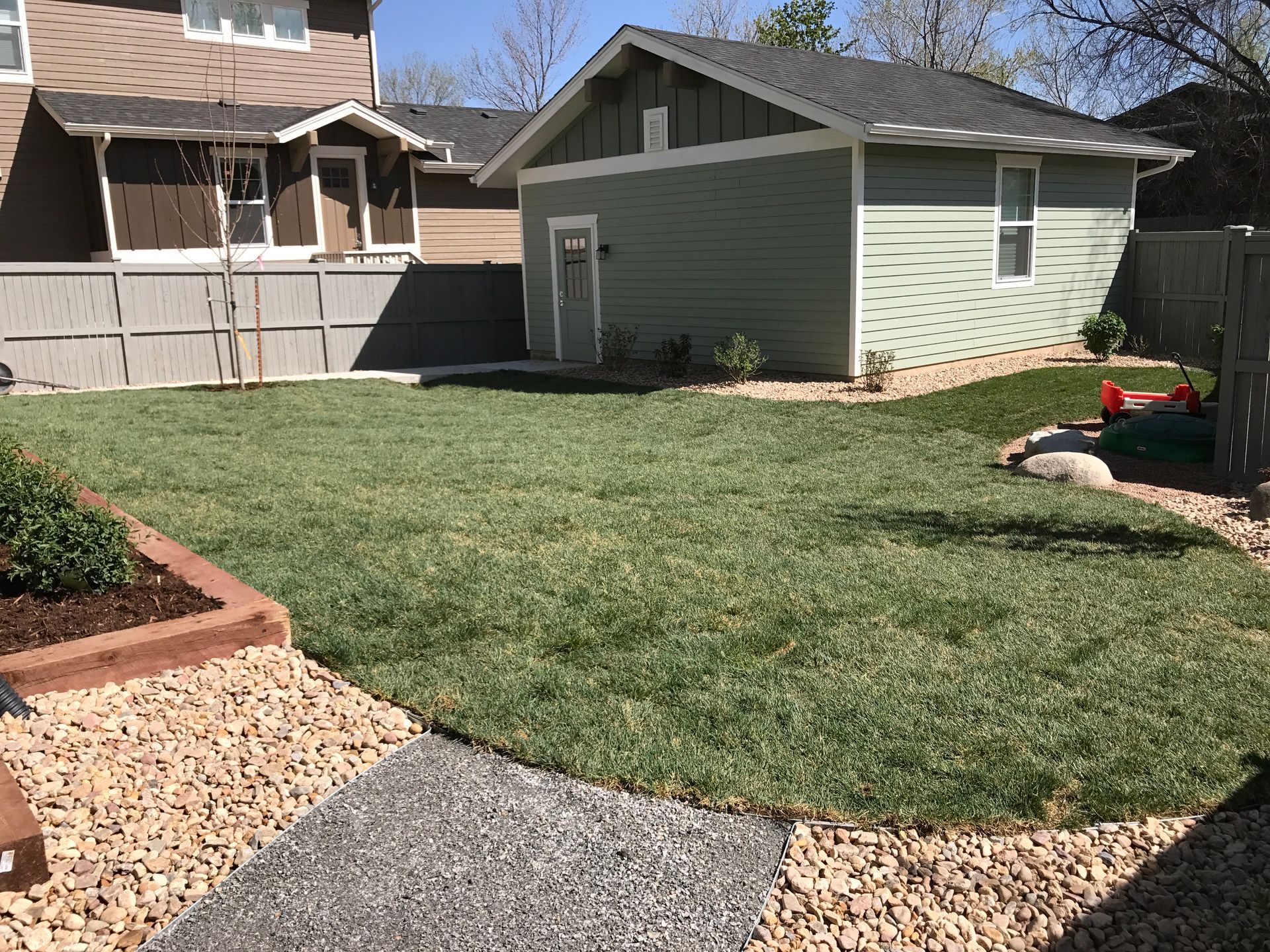 Lush green backyard with a small green shed, brown house, gravel path, and neatly trimmed lawn.