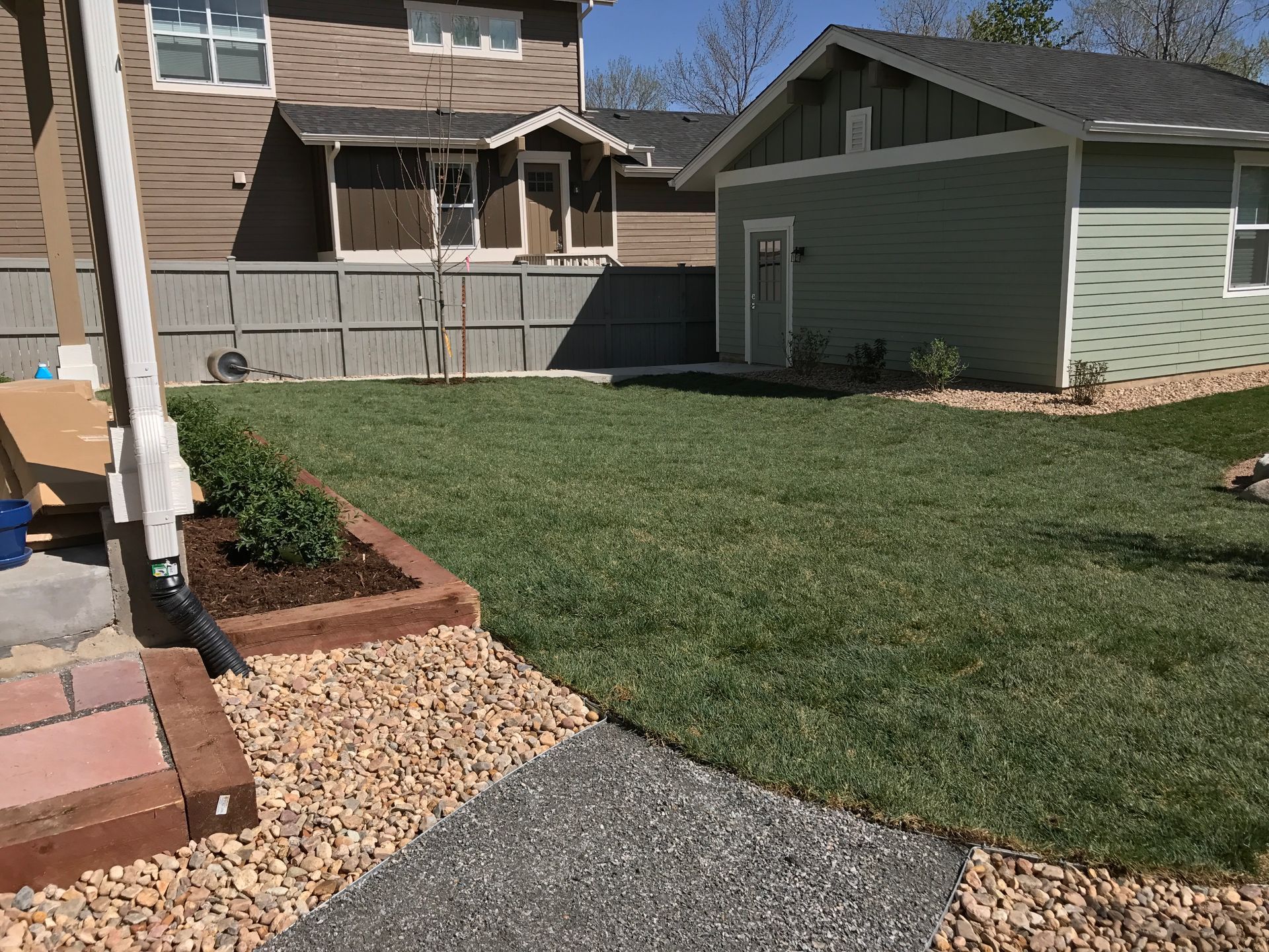 Backyard with green grass, gravel path, brown wooden border, small green shed, and houses in the background.