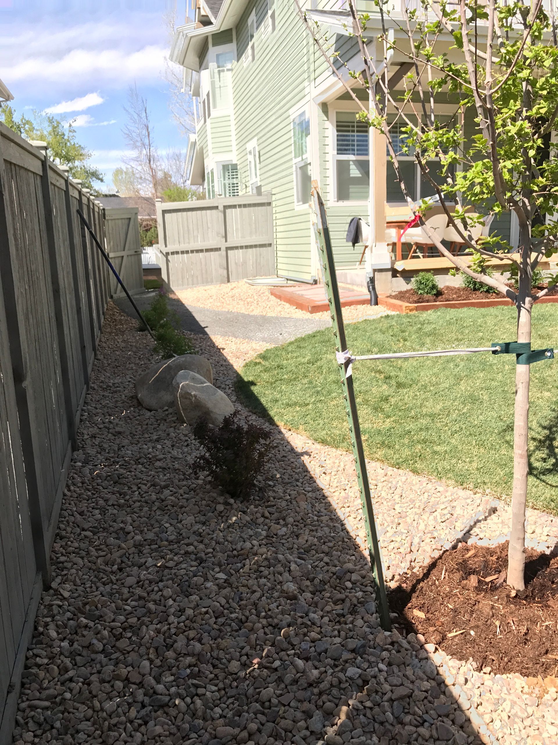 A narrow gravel side yard with a fence, large rock, and small tree, under a bright sky.