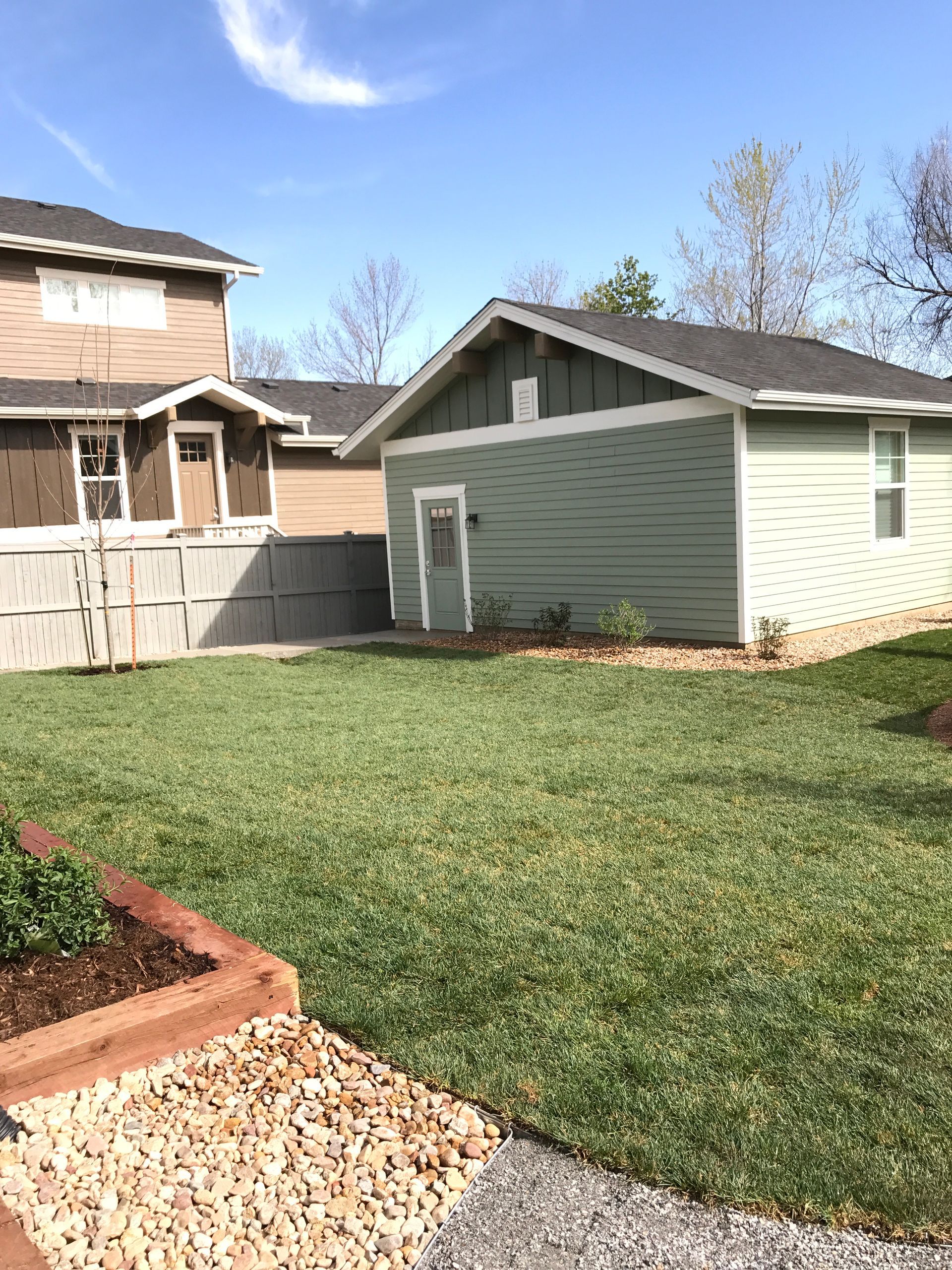 Green-sided building with dark roof, next to a building under construction. Lush green lawn with a stone path.