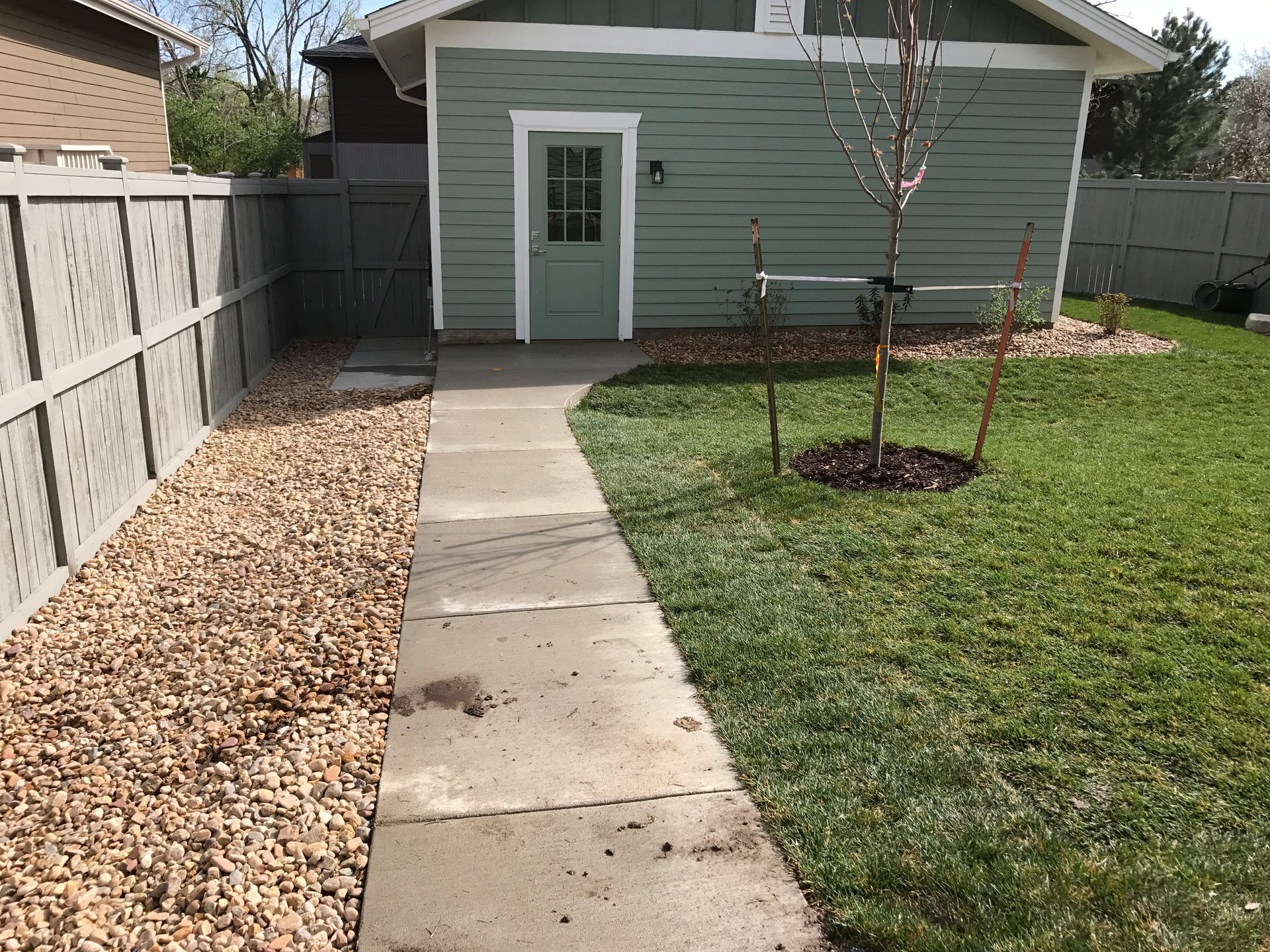 Concrete walkway leading to a light green shed with a door, flanked by a gravel bed and lawn.