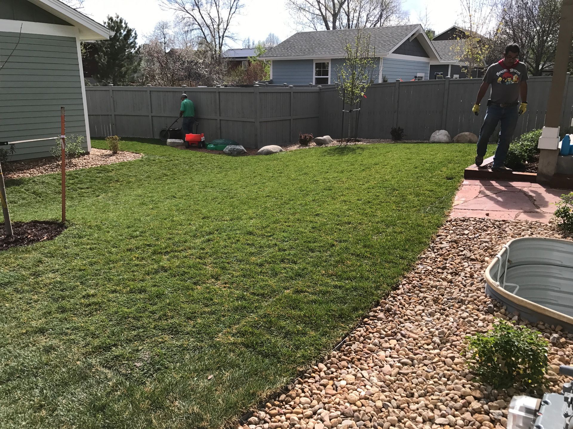 Man on patio watches as a person mows the green lawn in a backyard surrounded by a gray fence.