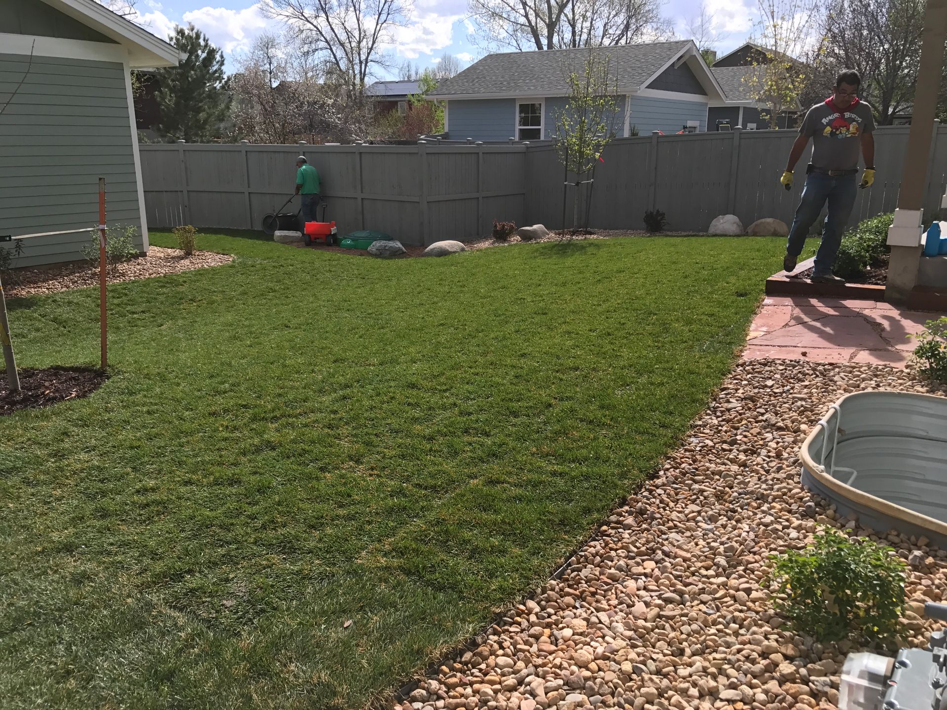 Man mowing a green lawn in a backyard; another man stands nearby. A gravel path and grey fence are also visible.