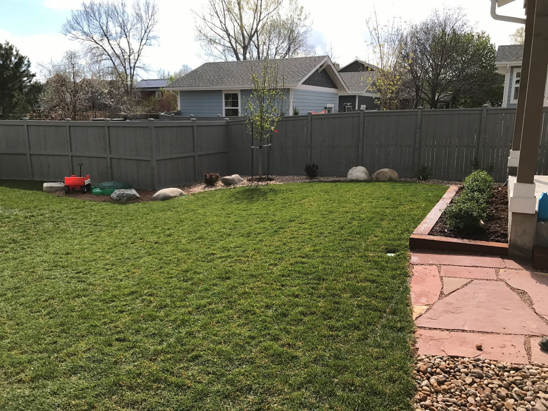 Green backyard with grass, a fence, small tree, and a stone patio.