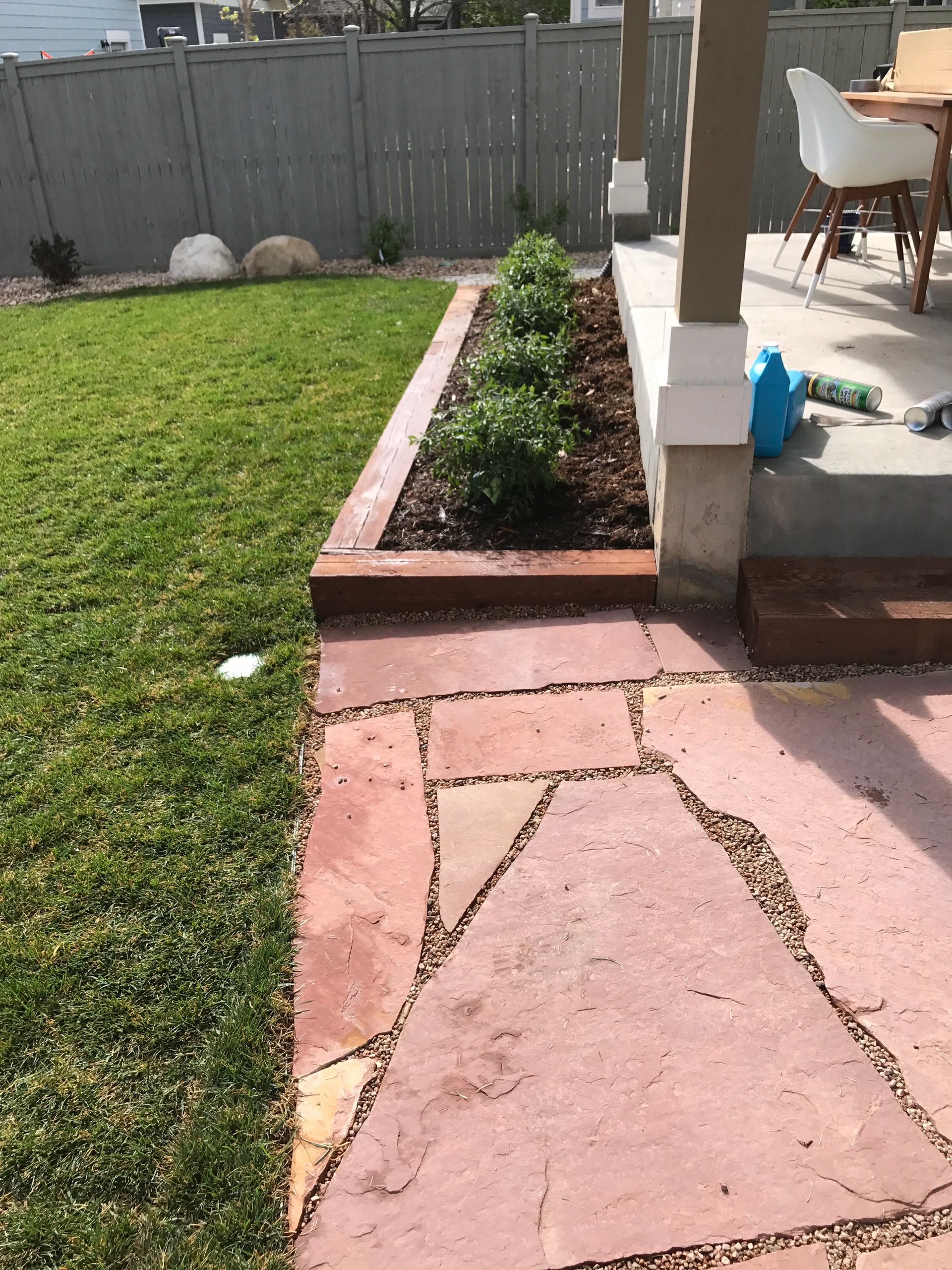 Stone path leads to a raised garden bed with plants, next to a grassy lawn and a patio.