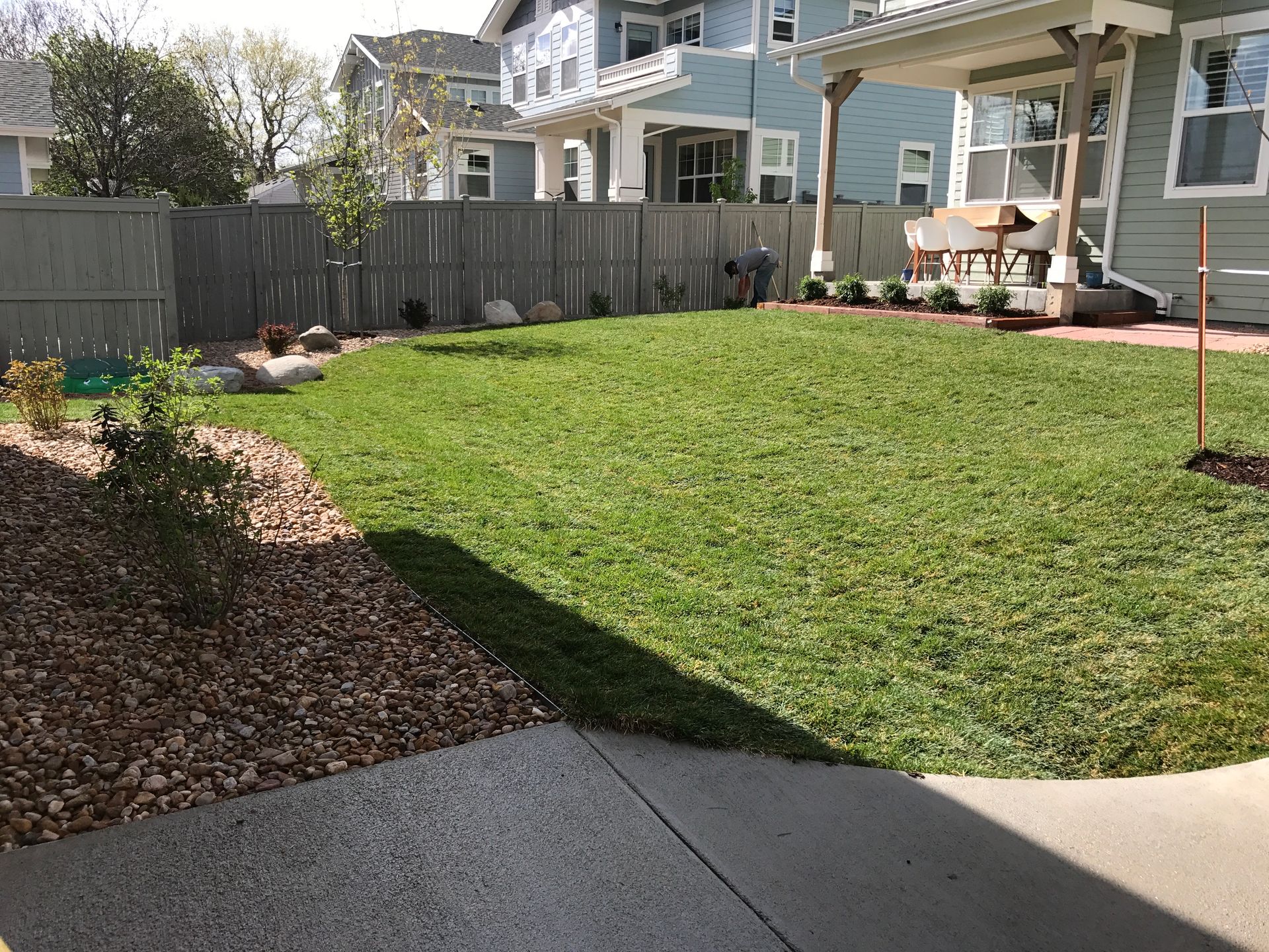 Lush green lawn with a gray fence and houses in the background. A concrete patio is in the foreground.
