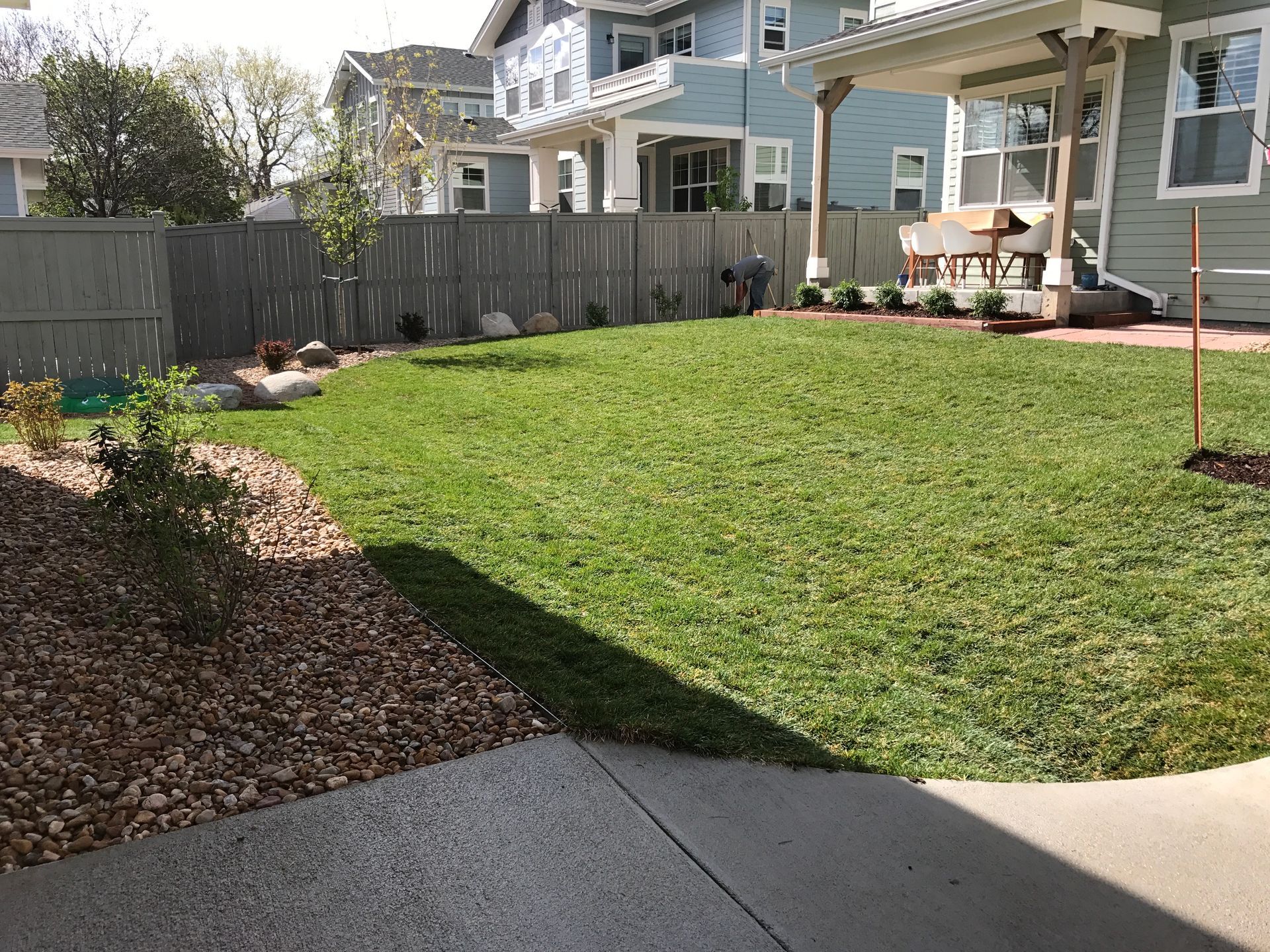 Backyard with green lawn, gray fence, blue houses, and a concrete patio.