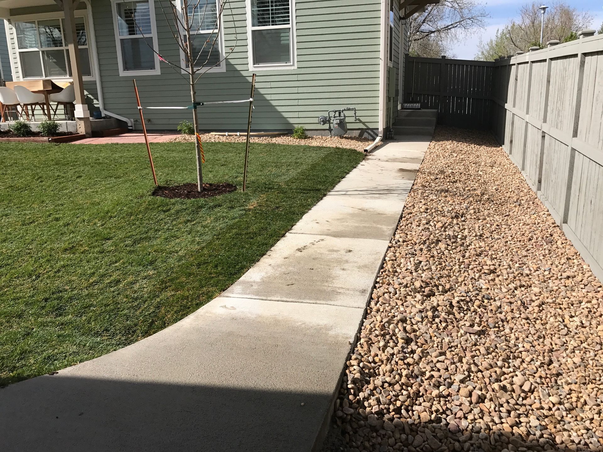 A concrete walkway curves past green grass and brown gravel beside a green house and gray fence.