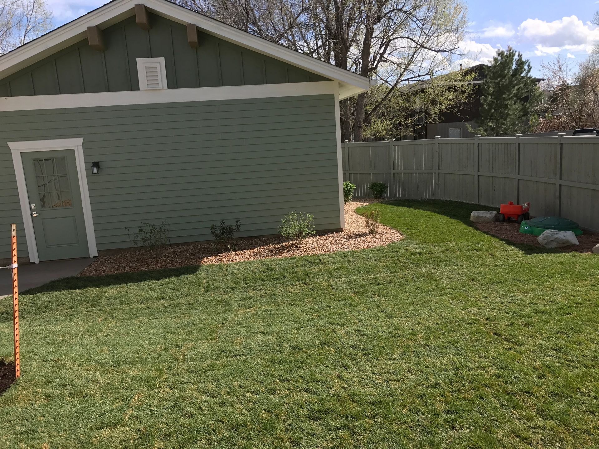 Green house and lawn with a wooden fence. A red play bin is on the grass.