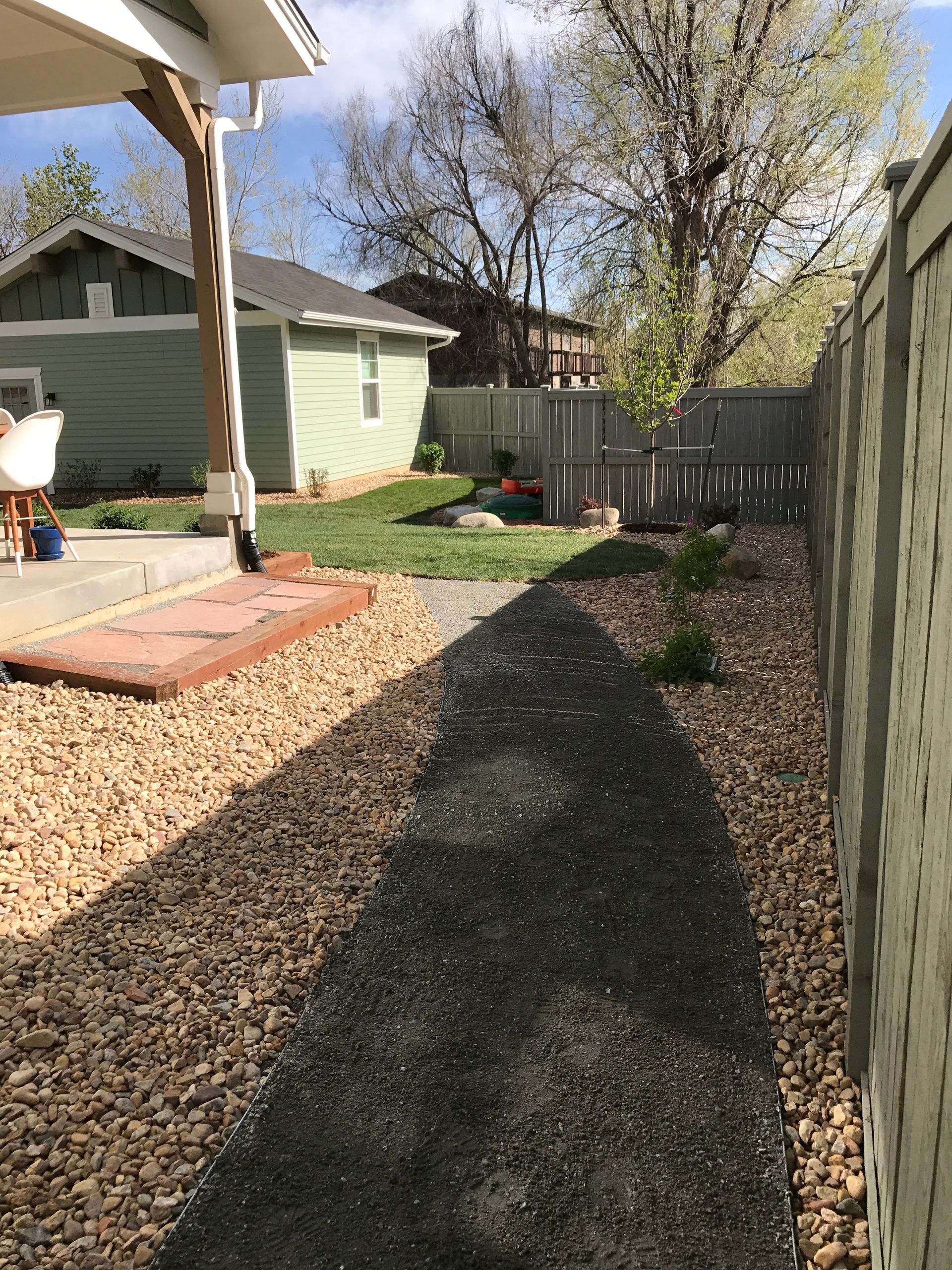 A gravel pathway bordered by rocks leads through a backyard, beside a fence and a small green building.