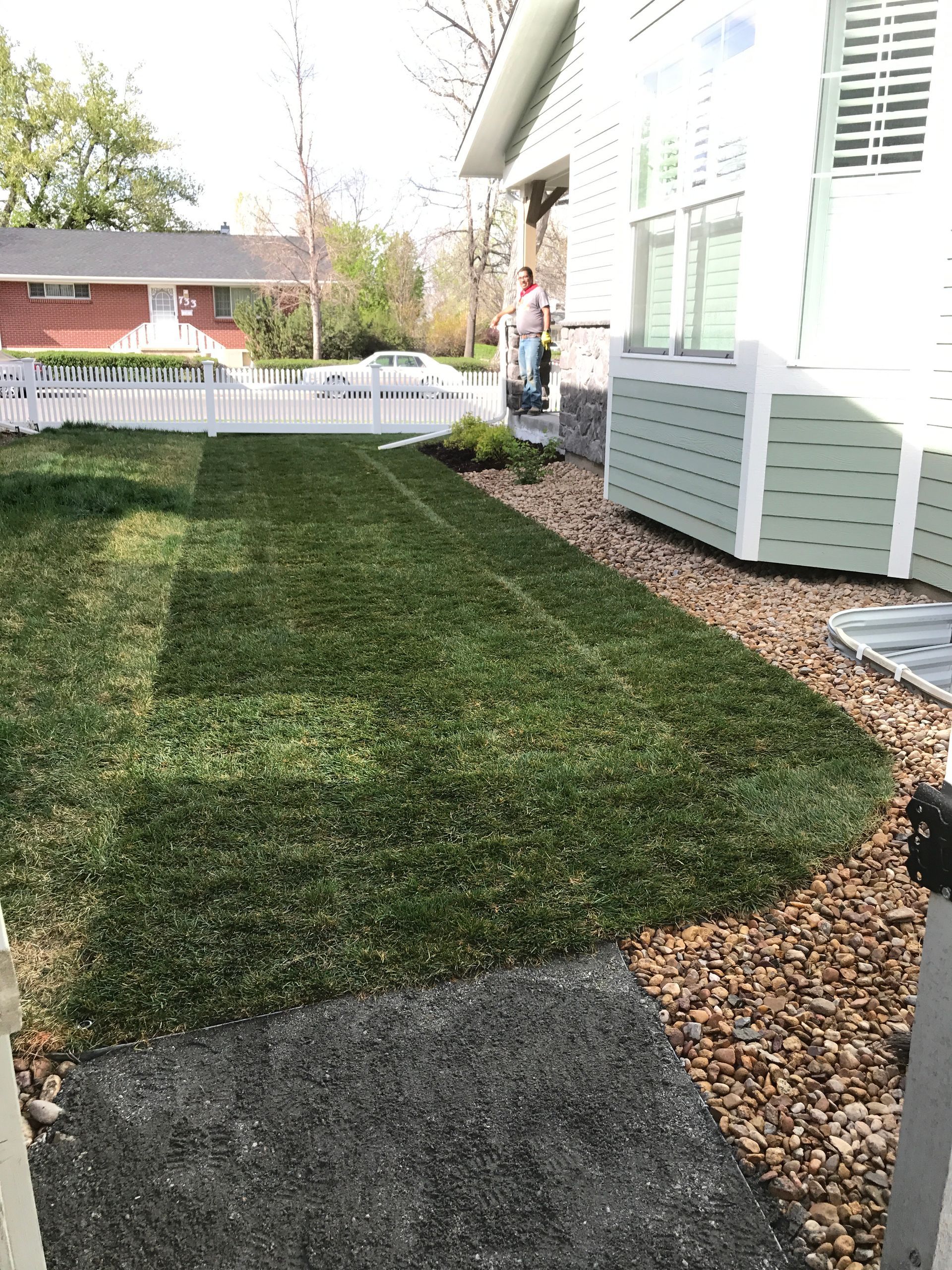 Lush green grass along a house with a gravel path, white picket fence, and a distant house.