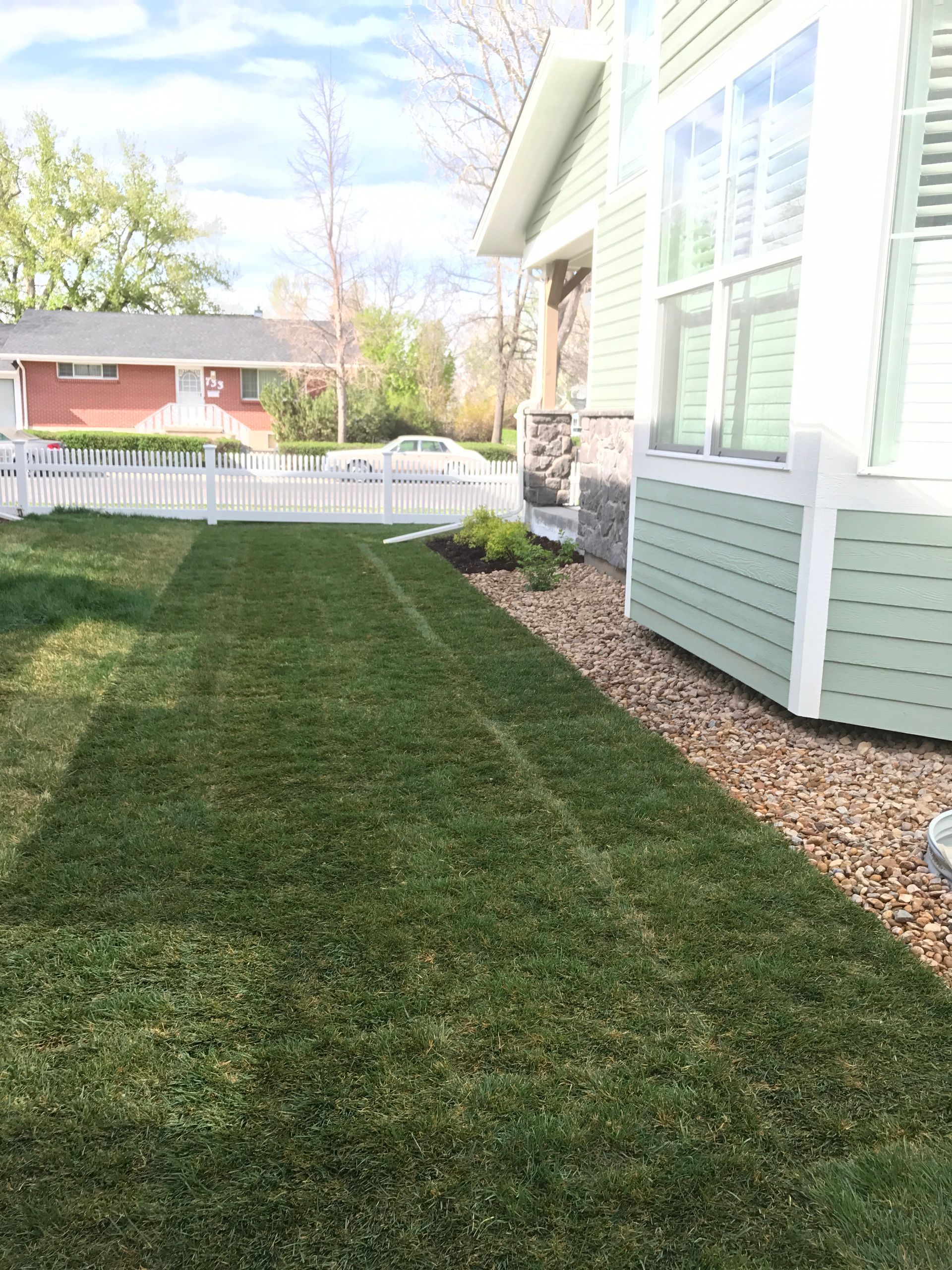 Green lawn next to a light green house with white trim and a small rock bed.