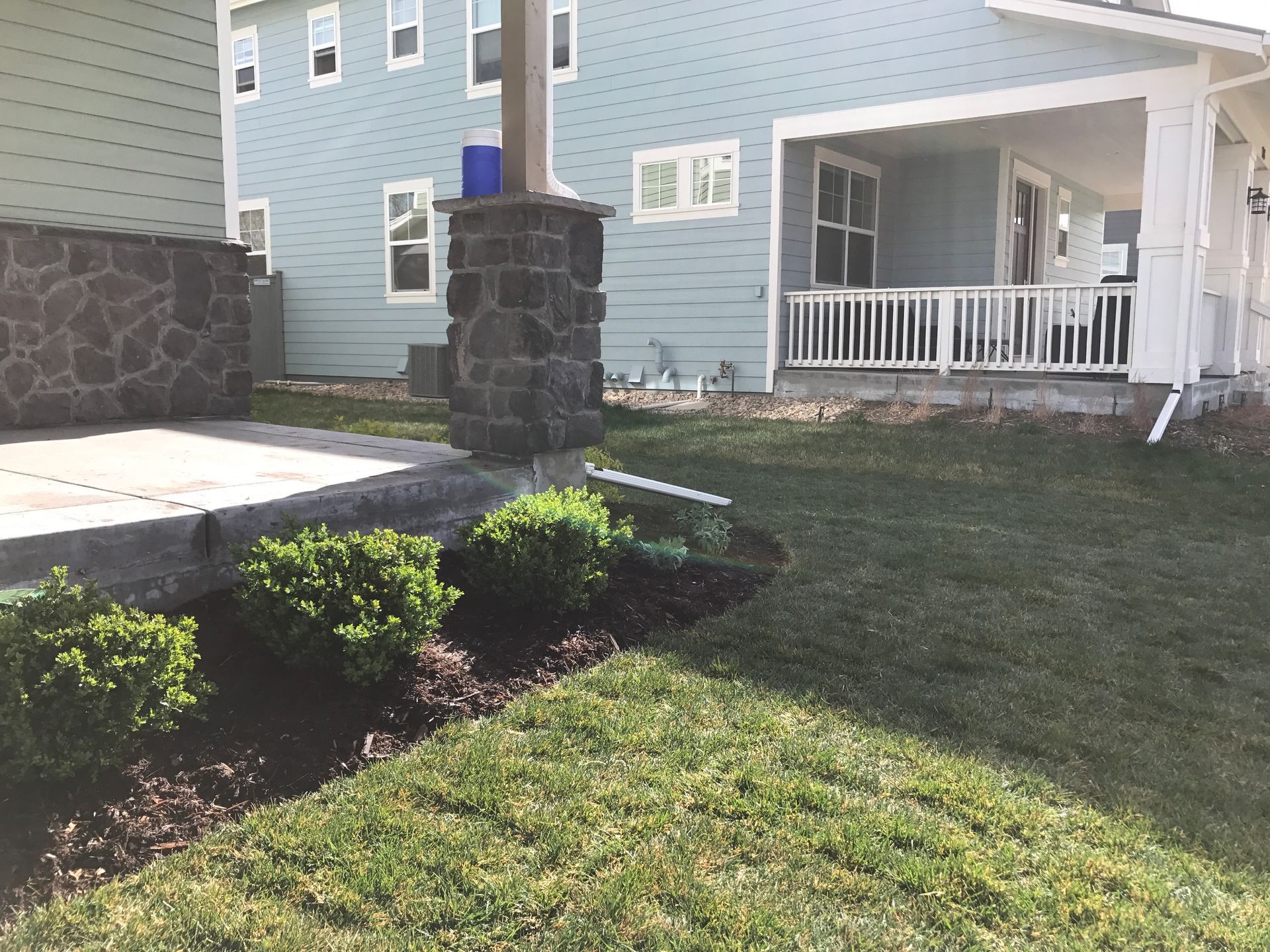Lawn and landscaping in front of light blue house with a porch and a column with stone siding.