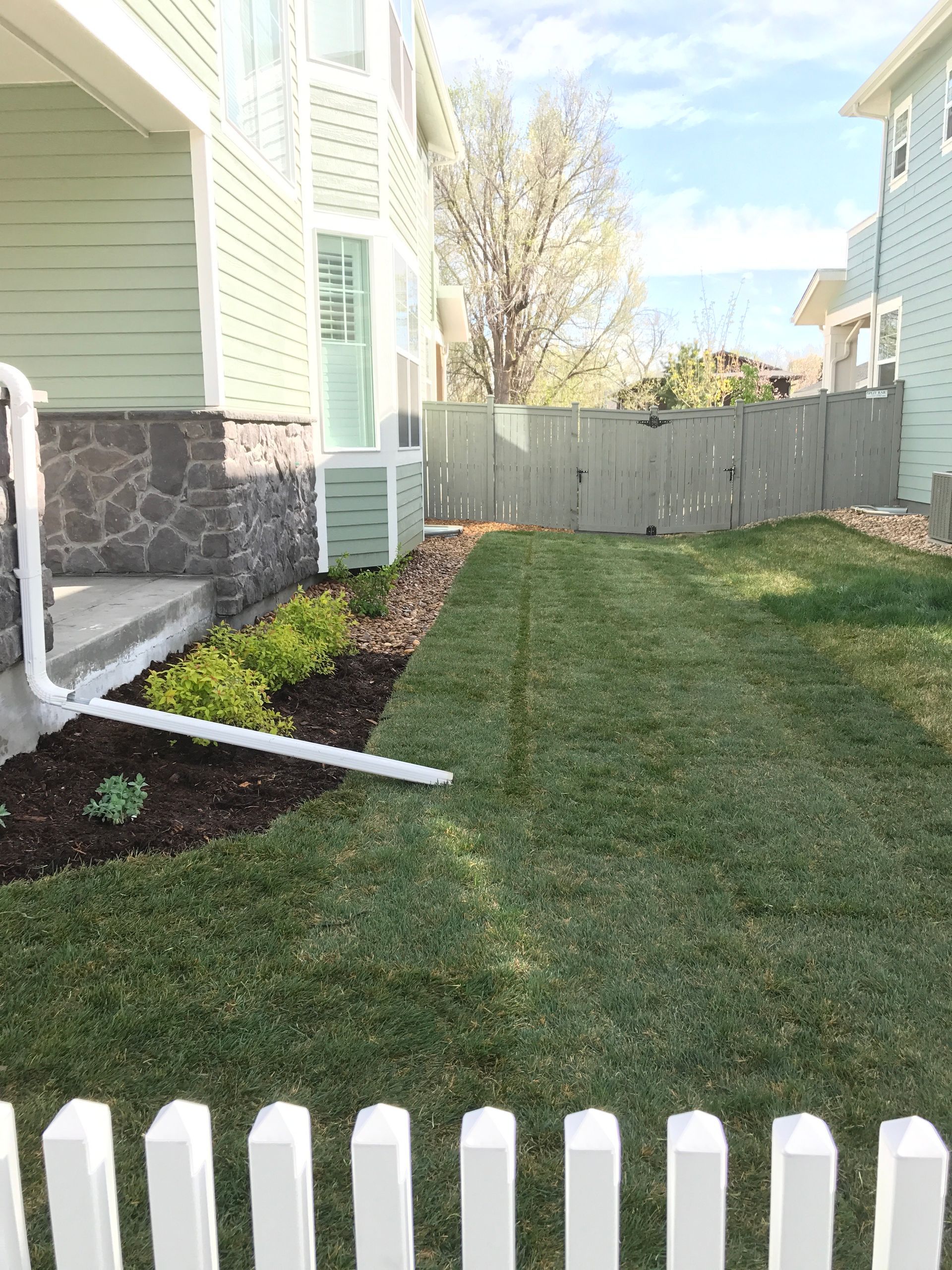 Green grass and garden next to a light green house with a white picket fence in front.