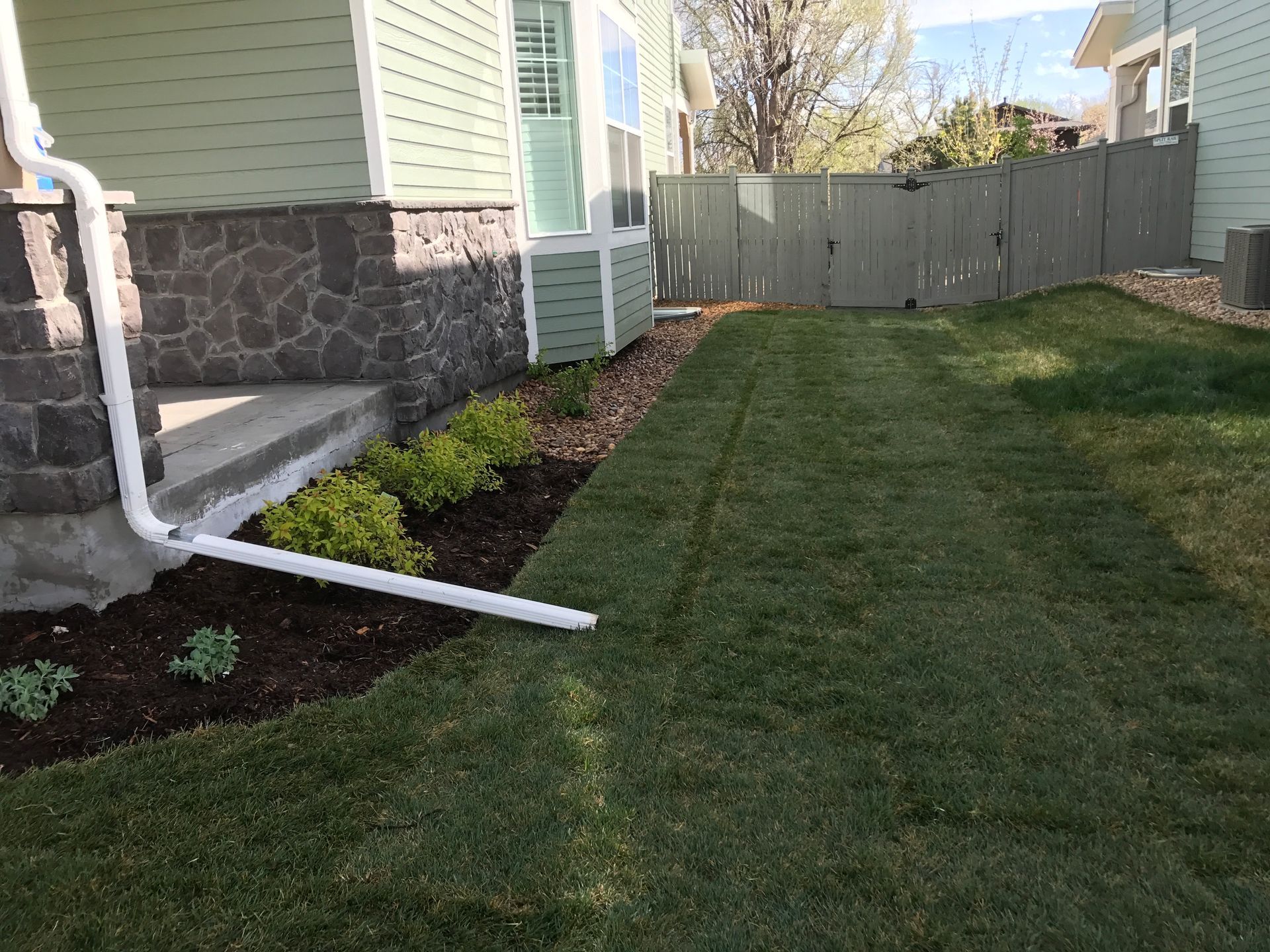 Green lawn bordering house with dark stone facade, white downspout, and wooden fence.