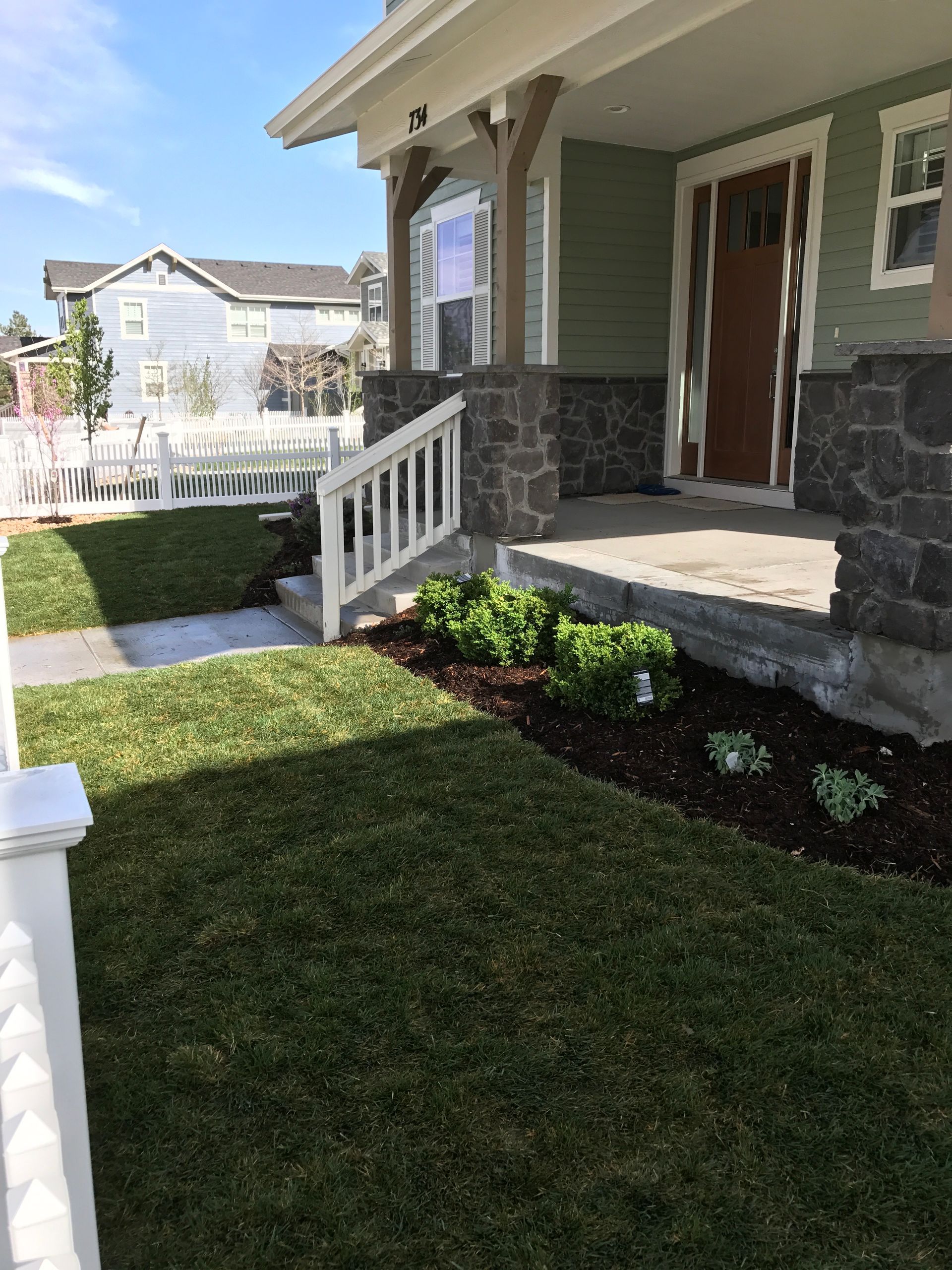 Front yard of a house with green lawn, white picket fence, and porch with plants.