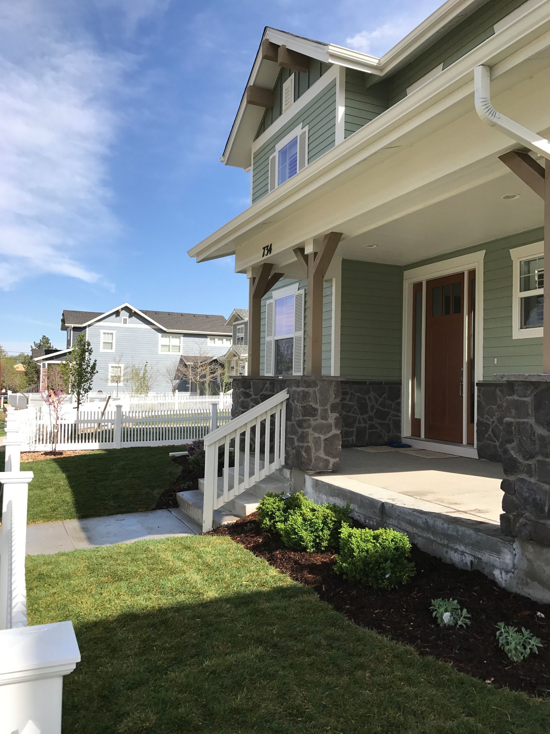 Green house with stone pillars, front porch, and white picket fence on a sunny day.