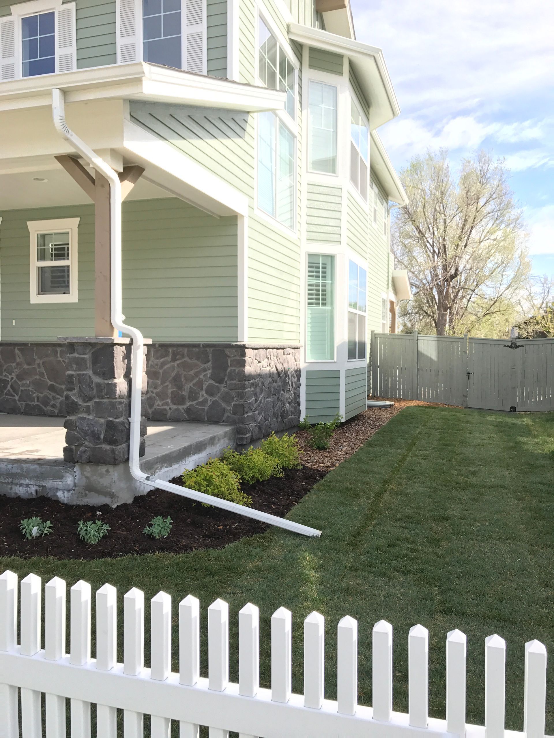 Green house with white trim, porch, and picket fence, next to a well-kept lawn and flower bed.
