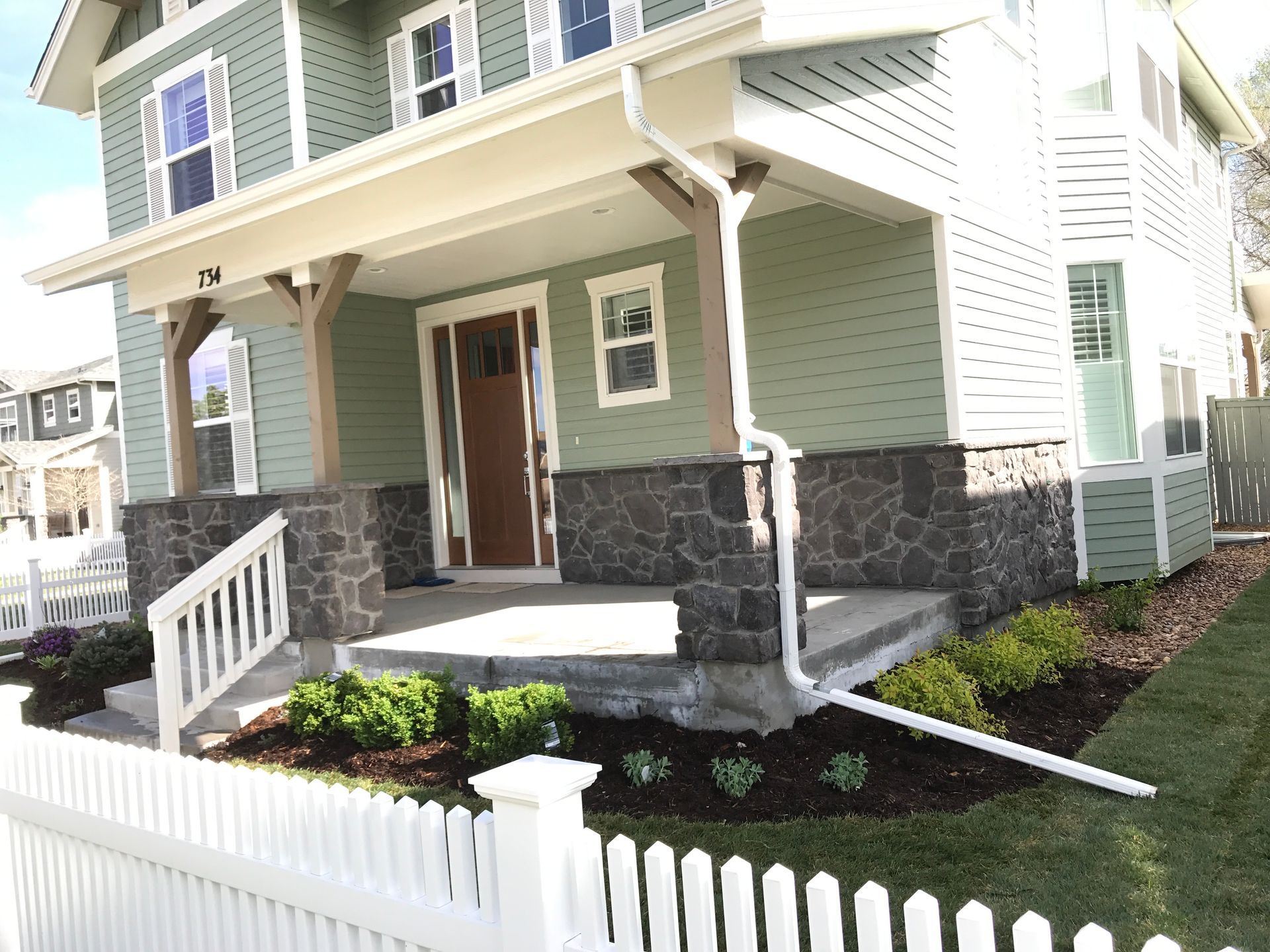 A two-story green house with stone accents, a porch, and a white picket fence in front.