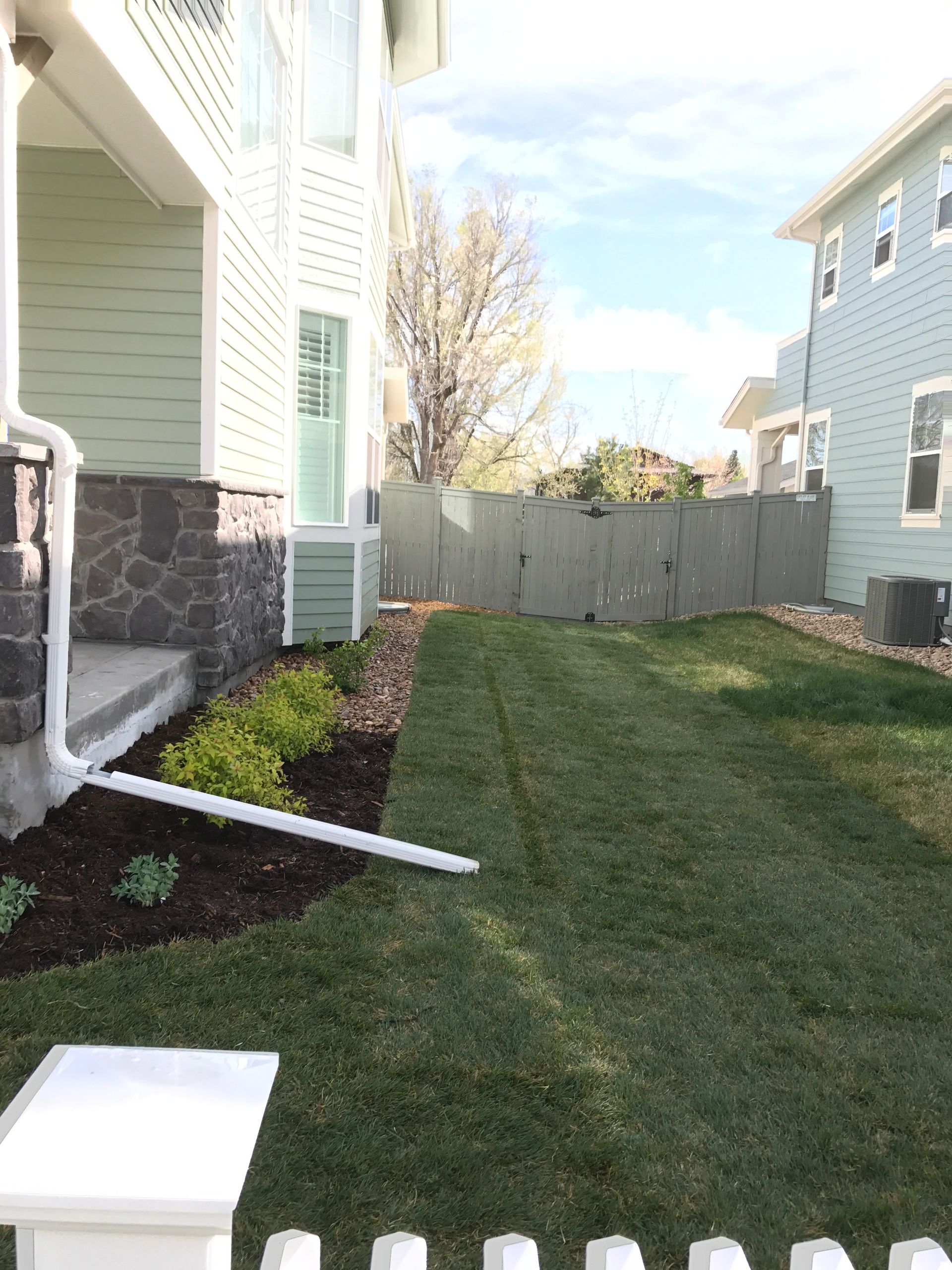 Lawn and house exterior. Green grass, mulch, and light-green house with stone accent. Fence in the background.