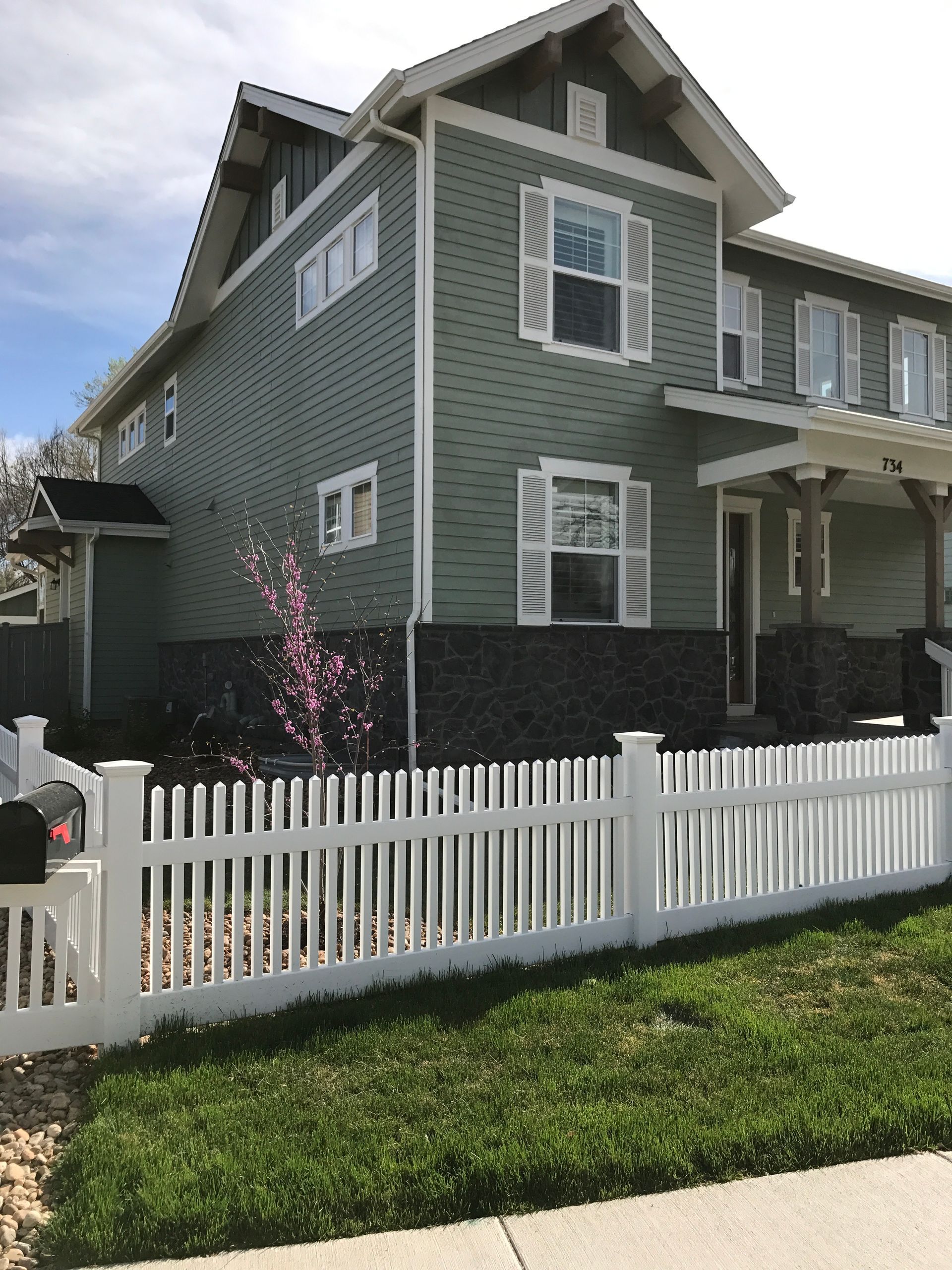 Two-story green house with white picket fence, a blossoming pink tree, and blue sky.