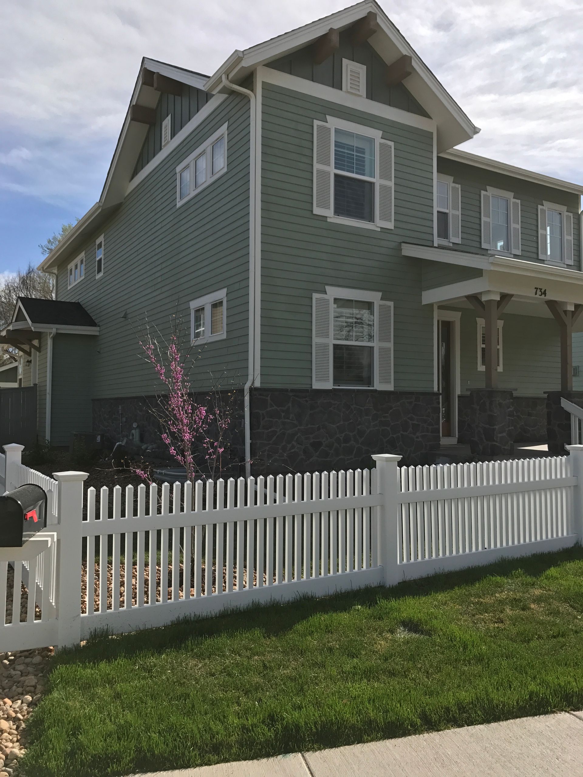 Two-story green house with white picket fence, shutters, and stone base. Sunny day.