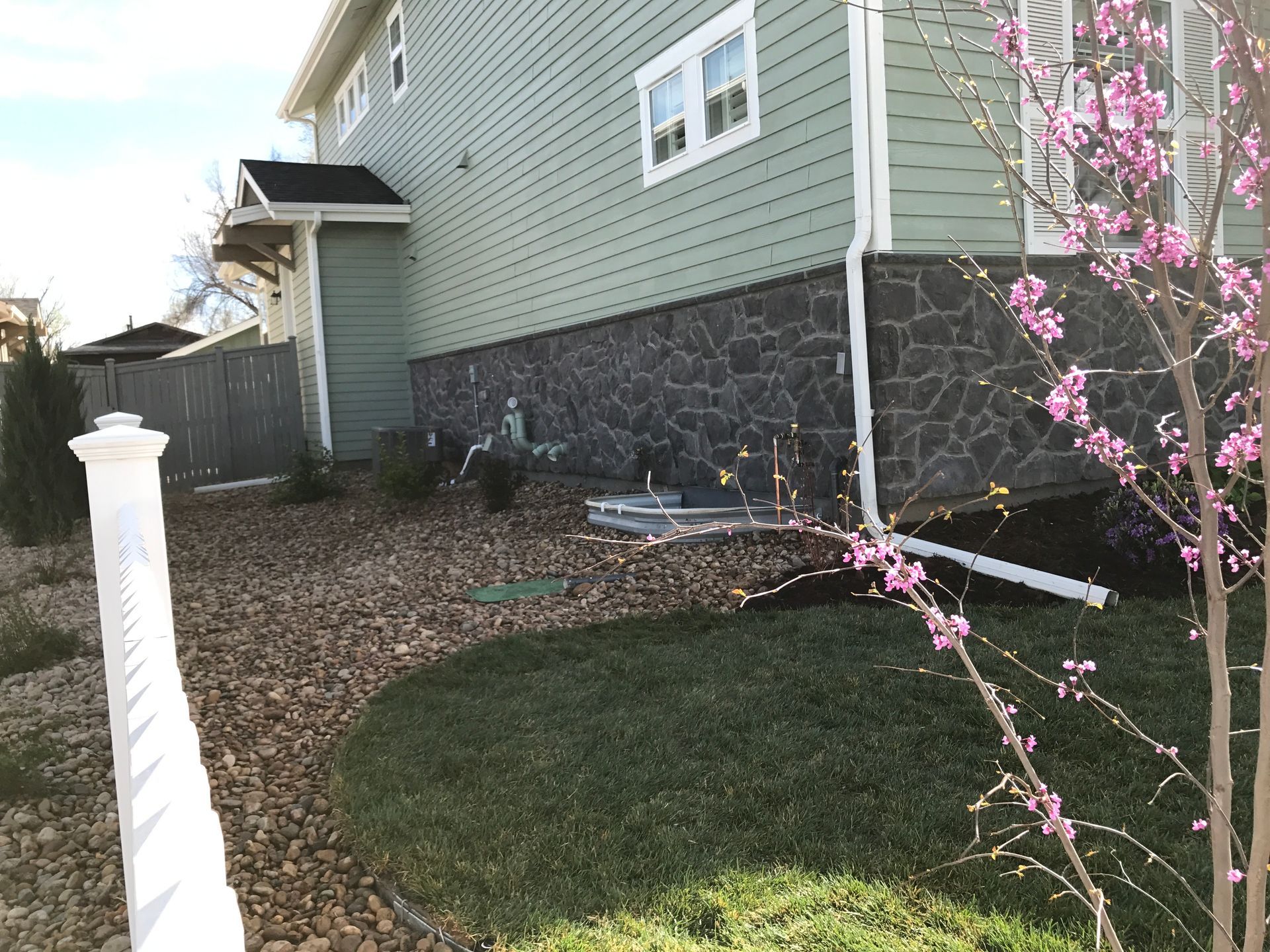 Side of a house with stone veneer, green siding, and a white fence, surrounded by landscaping.