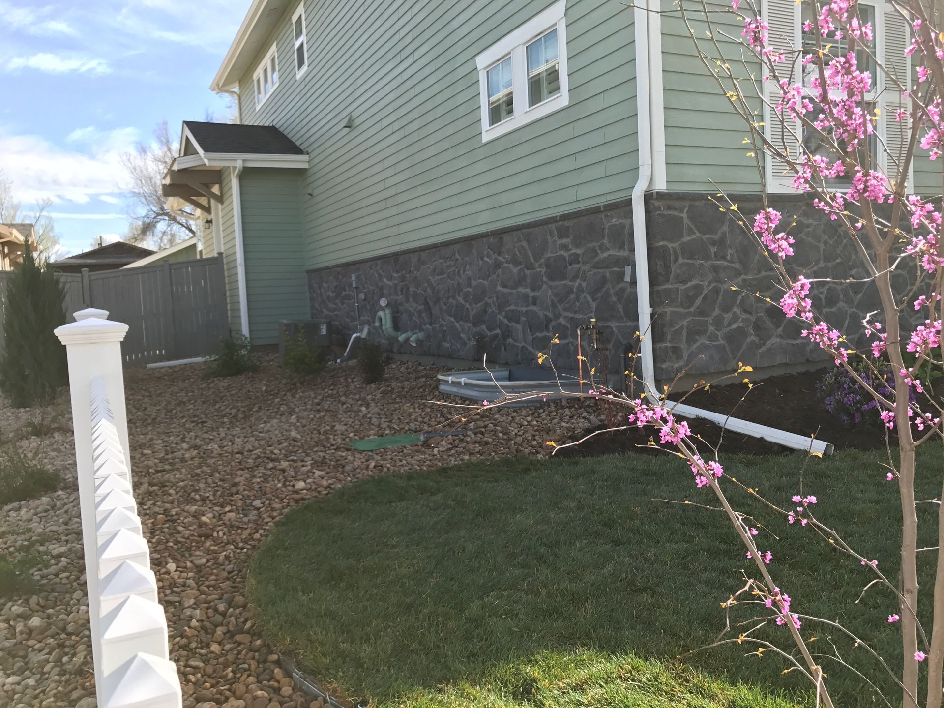 Side of a two-story green house with a dark stone foundation and a white fence next to a lawn and gravel.