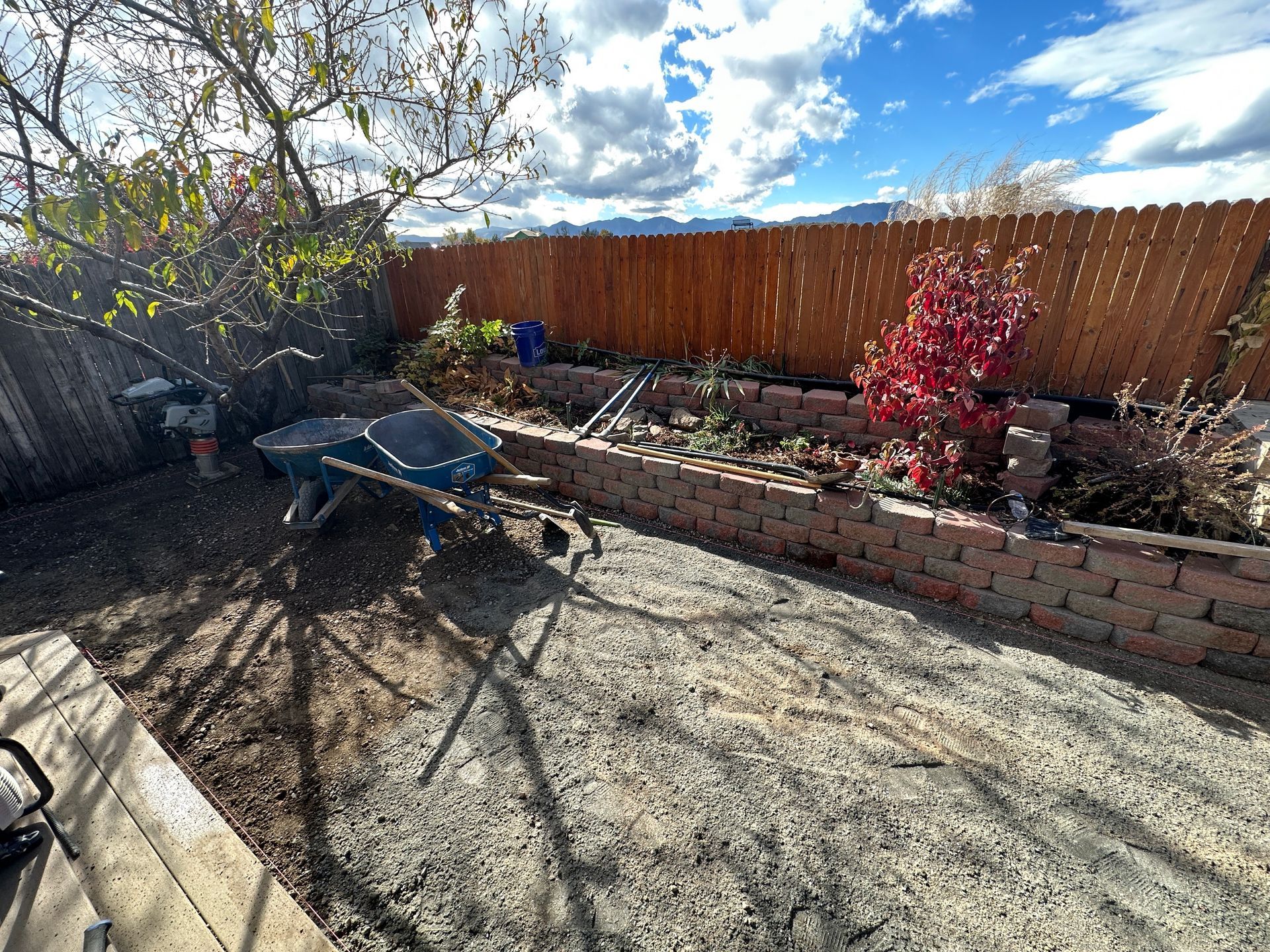 Backyard with wheelbarrow, brick planters, wooden fence, and blue sky.