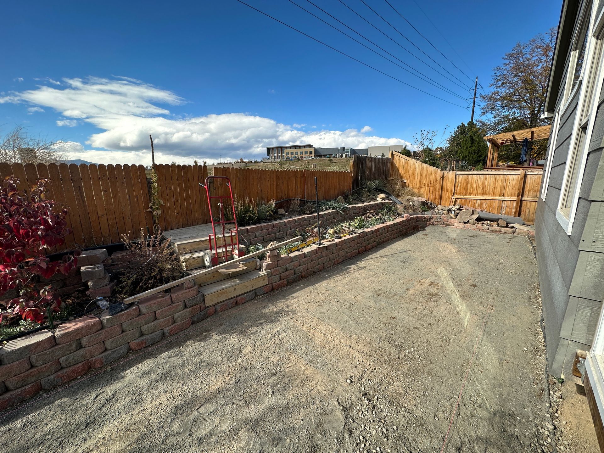 Backyard with gravel ground, a brick retaining wall, and a wooden fence under a blue sky.